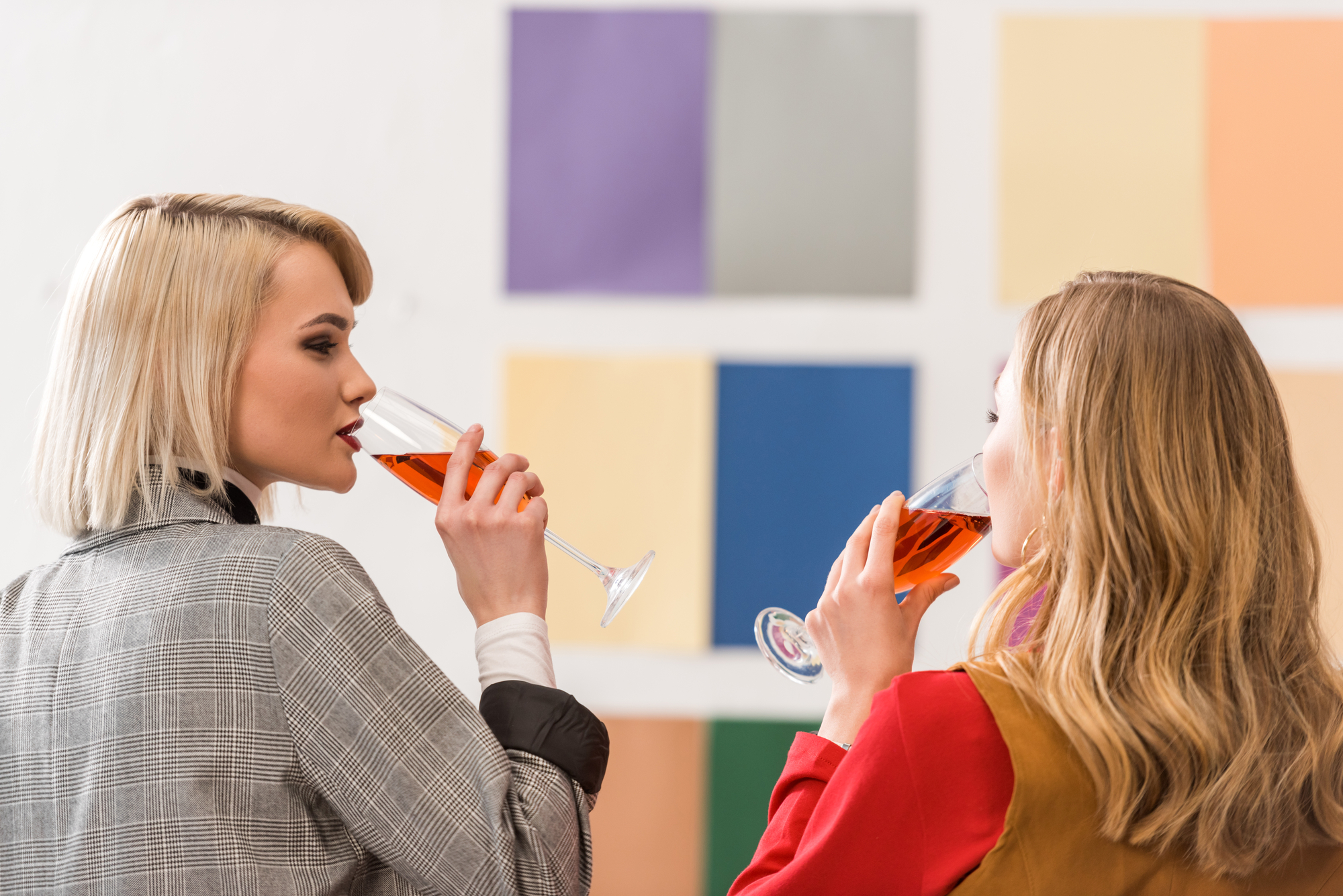 Two women, each with blonde hair, are standing and facing a colorful wall with square artworks. They are holding and sipping from wine glasses filled with a red beverage. One wears a checkered blazer, and the other wears a red top with a brown vest.