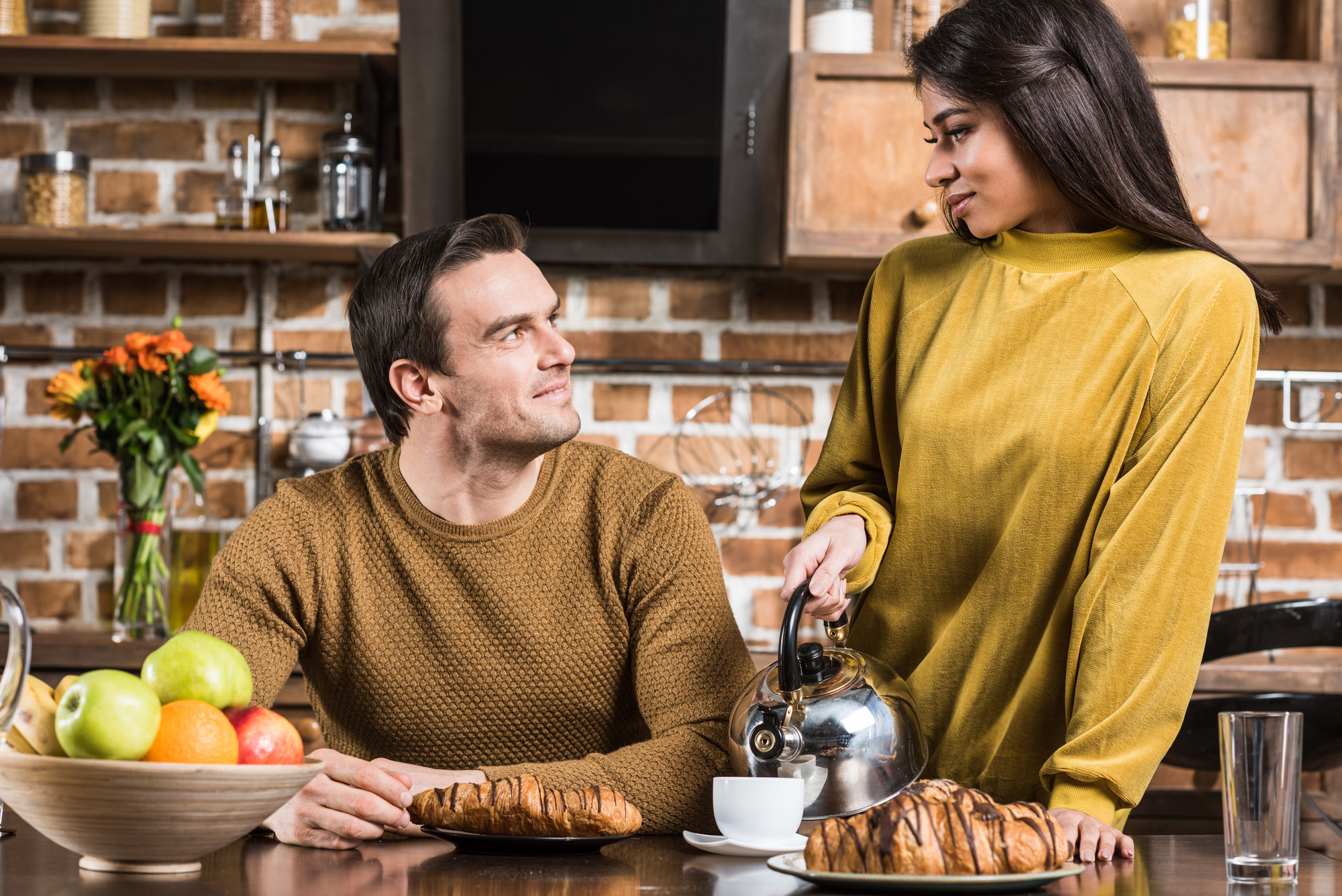 A man and woman are in a kitchen with a rustic brick wall. The man, seated at a table with croissants and a bowl of apples, is looking up at the woman, who is standing and pouring coffee from a kettle into a cup. Both are wearing cozy, earth-toned sweaters.