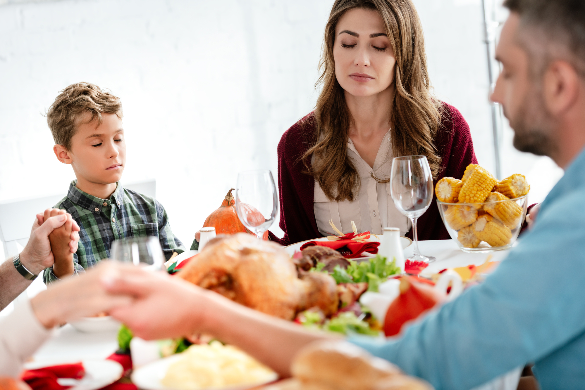 A family of four, consisting of two adults and two children, are sitting around a table holding hands and praying before a meal. The table is set with a roasted turkey, corn on the cob, and various vegetables. The scene appears to be during a holiday gathering.
