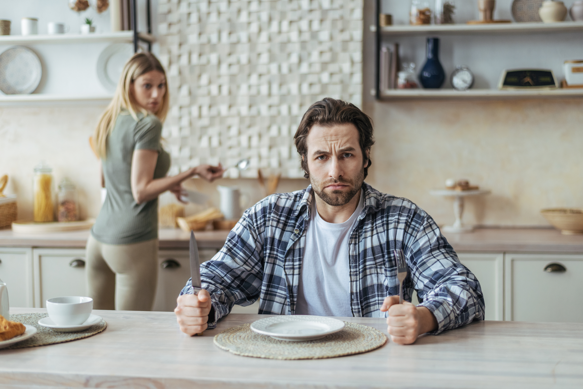 A man in a plaid shirt sits at a dining table with an empty plate, holding a fork and knife, looking frustrated. In the background, a woman in a green shirt prepares food at the kitchen counter and looks back towards the man. The kitchen is modern and well-lit.
