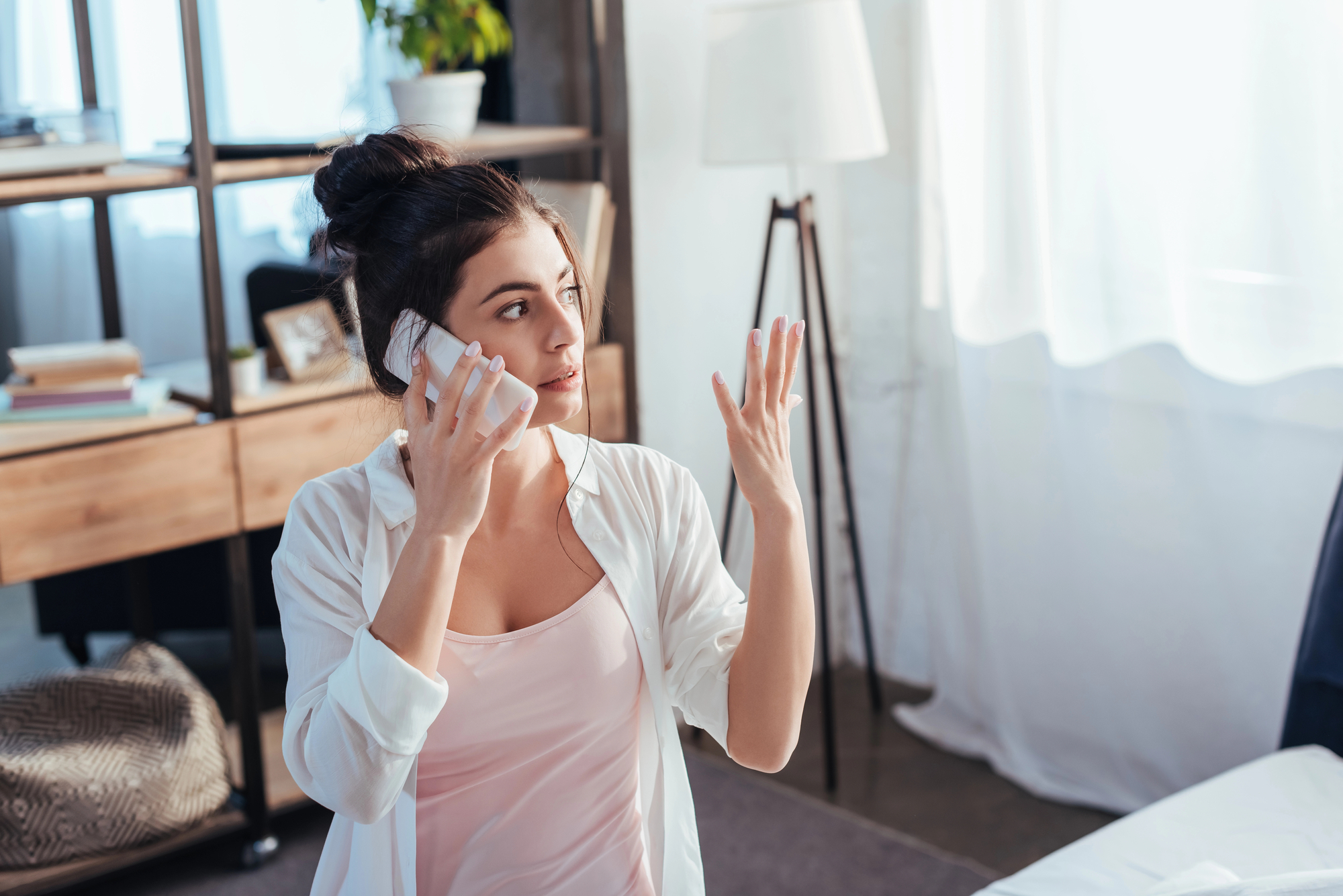 A woman with dark hair in a bun is sitting on a bed, holding a smartphone to her ear with one hand, and gesturing with the other. She is wearing a white shirt over a pink tank top and appears to be in a modern bedroom with bookshelves and a lamp in the background.