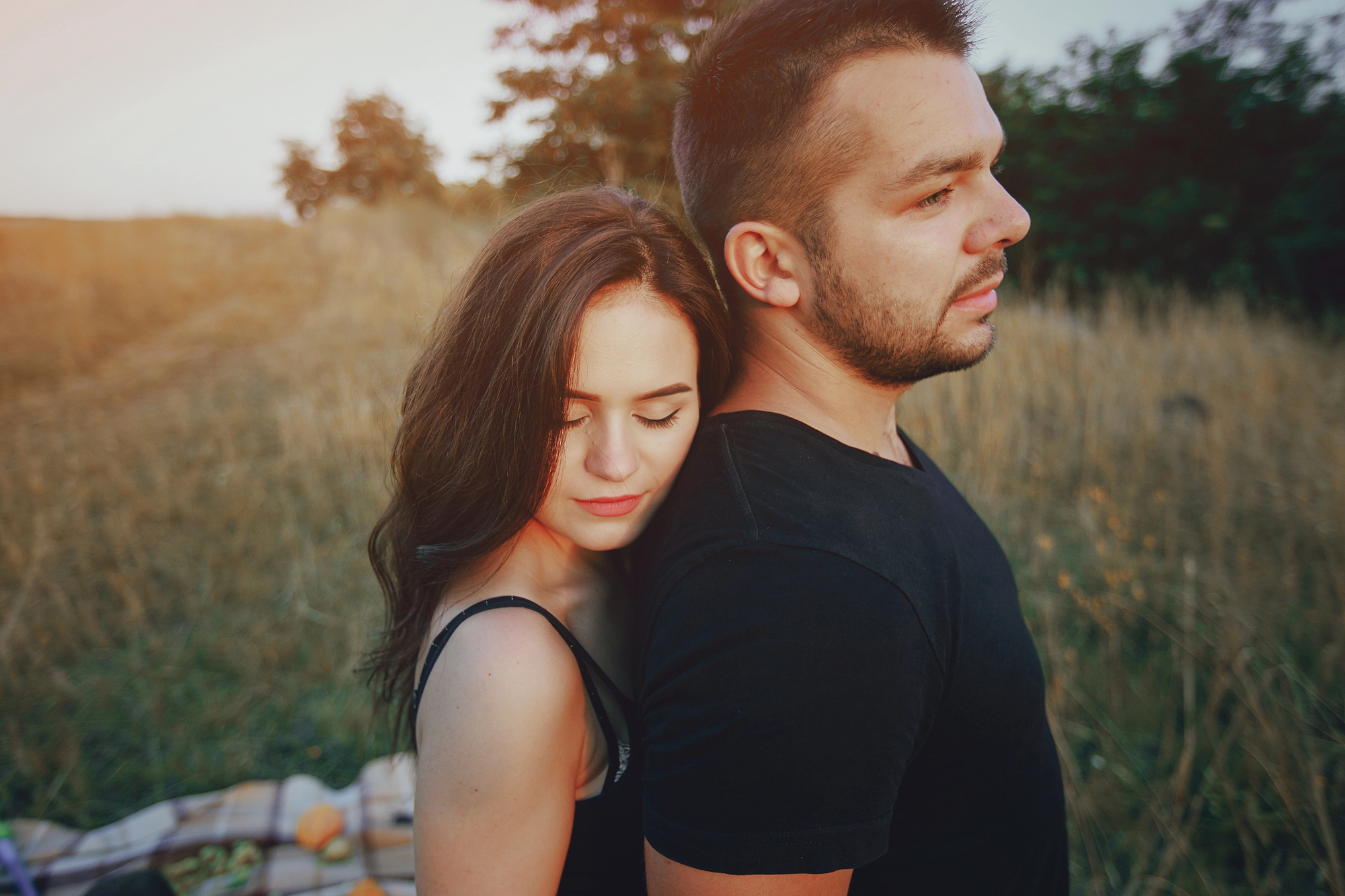 A woman with long dark hair and dressed in a black top leans her head on the back of a man with short dark hair, wearing a black T-shirt. They stand outdoors on a grassy field during sunset, with trees in the background and a blanket with items on the ground.