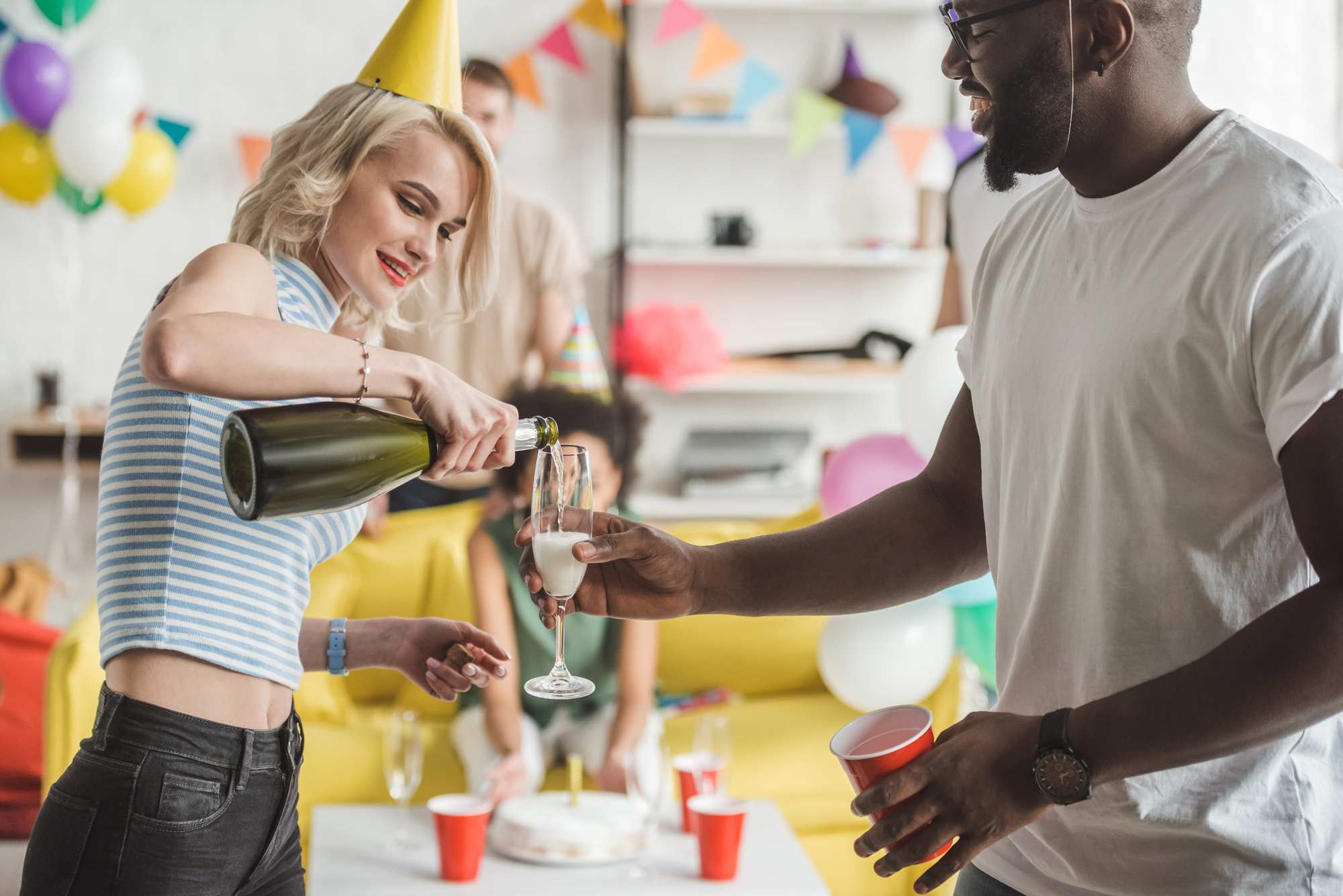 A woman in a party hat is pouring champagne into a glass held by a man at a festive gathering. They stand in a colorful room decorated with balloons and bunting, with other people and party supplies visible in the background. Both appear to be smiling and enjoying the moment.