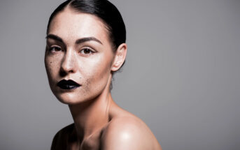 A woman with dark hair slicked back is posing against a neutral gray background. She has prominent freckles, dark bold eyebrows, and is wearing glossy black lipstick. Her shoulders are bare, and she has a serious expression.