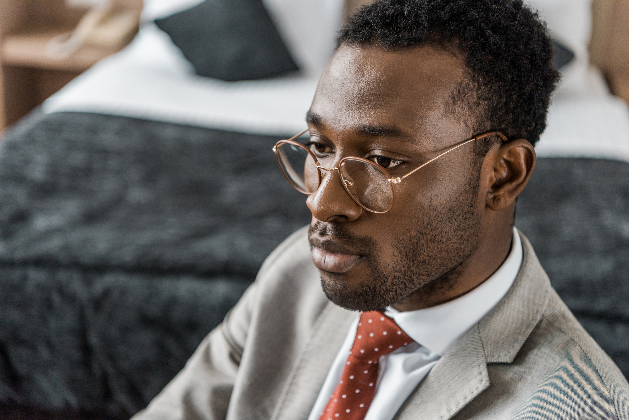 A man with short curly hair and glasses is seen in a close-up portrait. He is wearing a light gray suit jacket, white dress shirt, and a red tie with white polka dots. The background is slightly blurred, showing a bed with pillows in a tidy room.