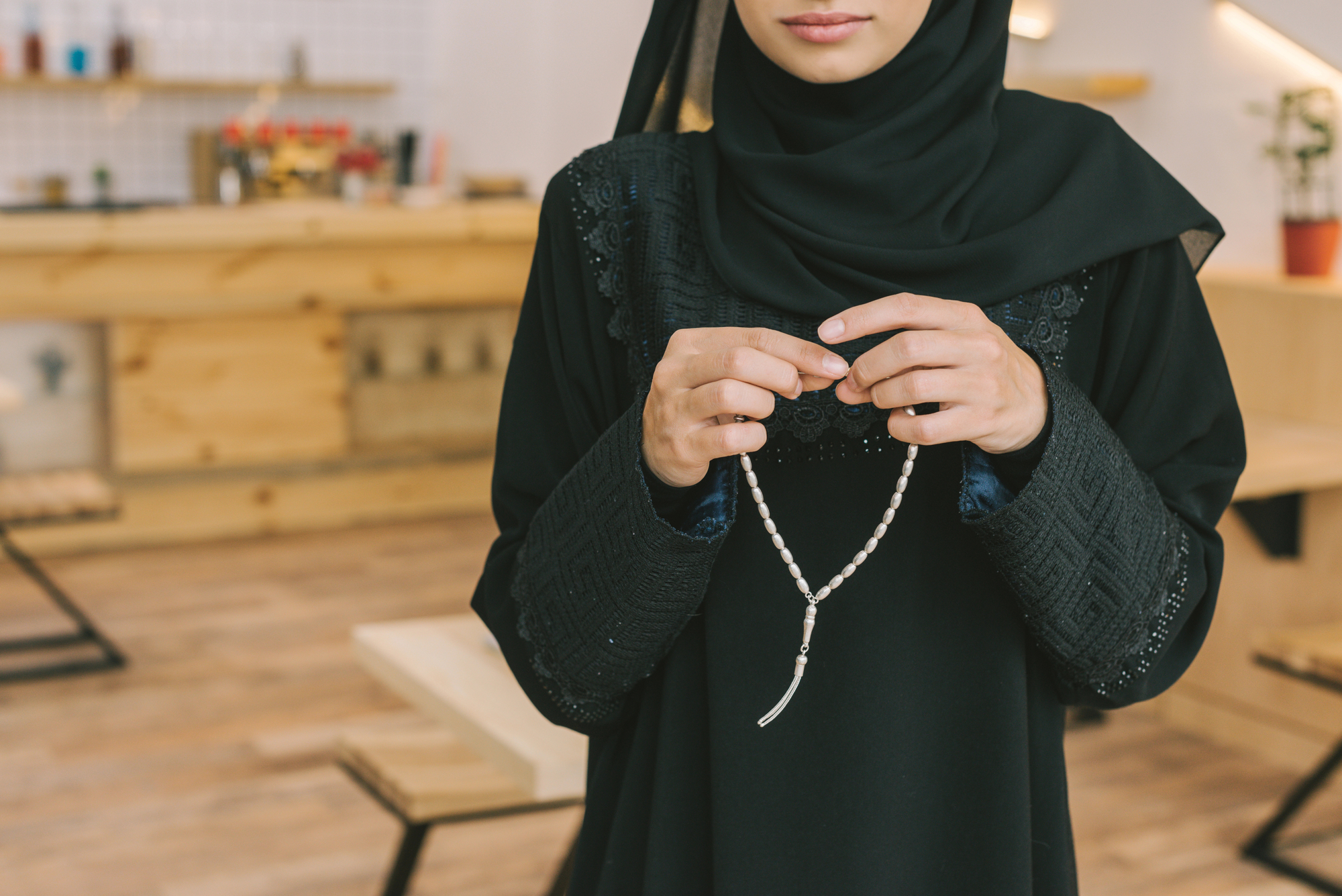 A woman dressed in a black abaya and hijab holds a string of prayer beads in her hands. She is standing inside a modern, light-colored room with wooden furniture and a small potted plant in the background. The focus is on her upper body, hands, and prayer beads.