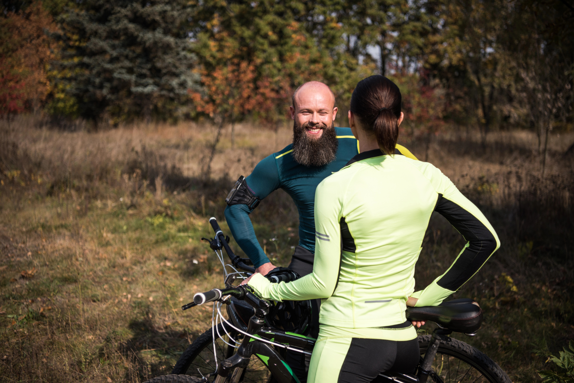 Two cyclists take a break on a trail surrounded by autumn foliage. A bearded man in teal cycling gear smiles while straddling his bike, engaging in conversation with a woman in bright yellow and black cycling attire who stands beside her bike.