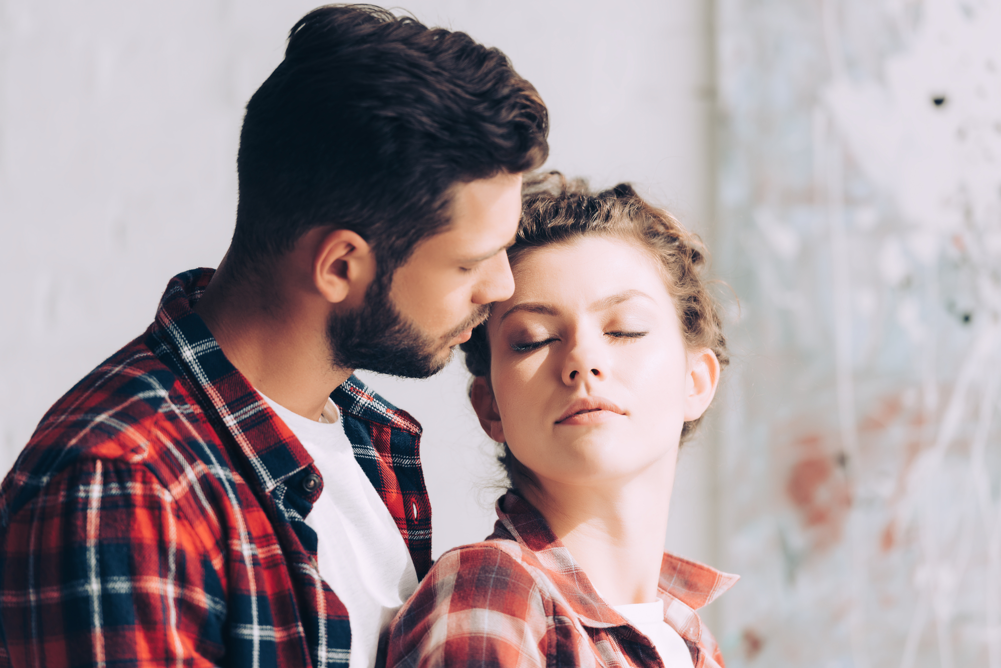 A man and woman stand closely together. The man, with a beard and short hair, looks down at the woman while gently touching his forehead to hers. The woman, with her eyes closed and relaxed expression, slightly leans into the man. Both wear red plaid shirts.
