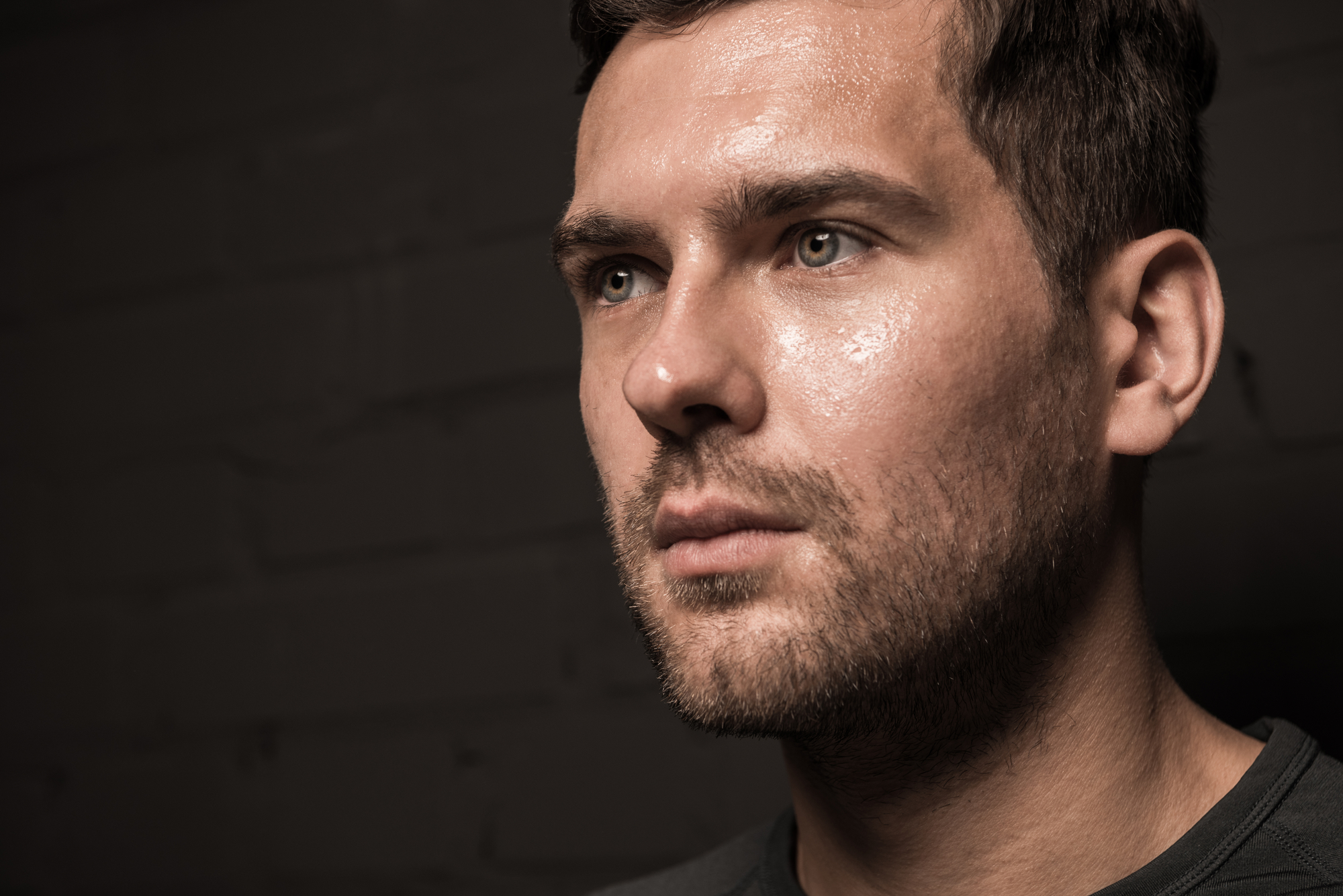 A close-up of a man with short dark hair and a beard, looking slightly to the side. The background is dark, with subtle shadows highlighting his facial features. His expression appears thoughtful and focused.