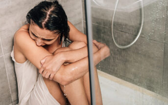 A woman sits on the floor in a shower, holding her knees close to her chest. She looks distressed and wet from the running water, which is visible from the showerhead. The setting appears to be a modern, tiled shower.