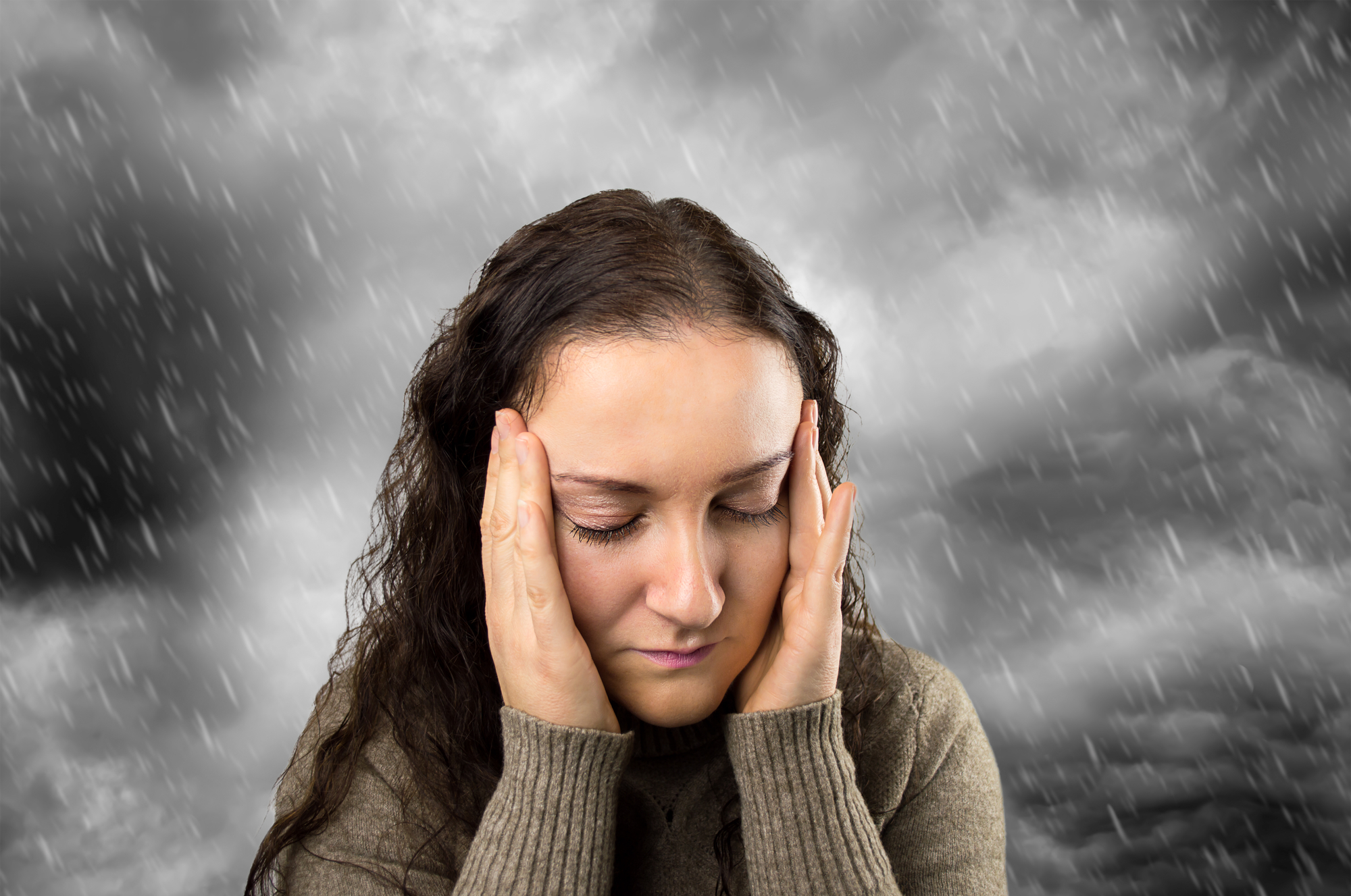 A woman with closed eyes and a distressed expression holds her hands to her head. She is wearing a brown sweater. Behind her, a dramatic black-and-white stormy sky with heavy rain emphasizes the intensity of her emotion.