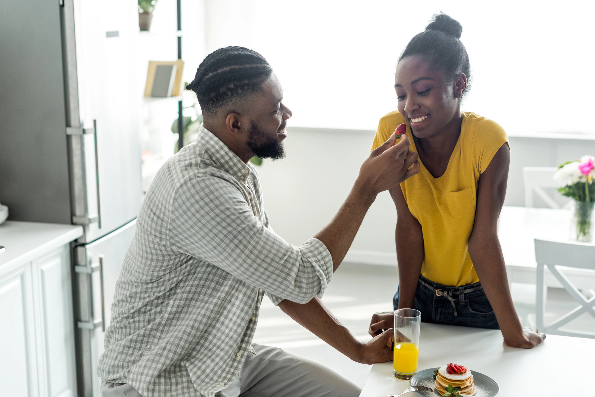 A man is feeding a woman a piece of fruit while she leans on a kitchen counter, smiling. On the counter, there's a plate with pancakes and a glass of orange juice. The scene is bright and cheerful, with flowers and a refrigerator in the background.