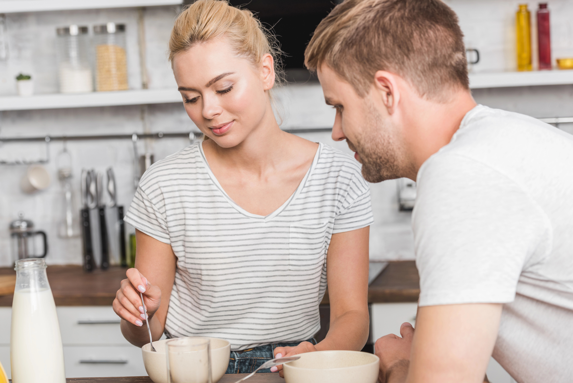 A woman with blonde hair wearing a striped shirt stirs a bowl with a spoon while a man with short brown hair, also in a white shirt, looks on. They are sitting at a kitchen counter with a bottle of milk and another bowl nearby.