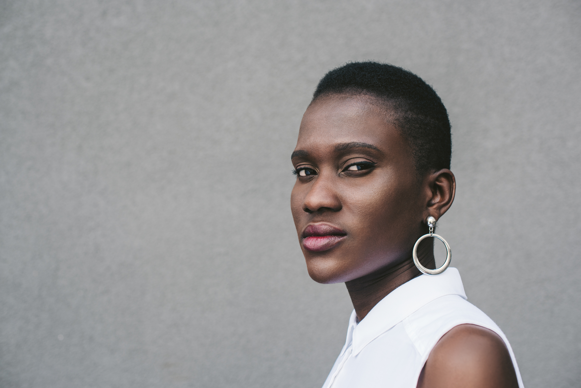 A person with short hair and hoop earrings gazes confidently towards the camera. They wear a white sleeveless top against a plain grey background. Their expression is calm and self-assured.
