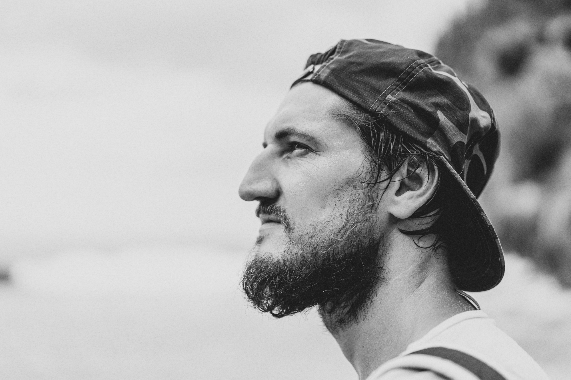Black and white close-up of a man in profile, wearing a backwards baseball cap, with a beard and focused expression. He appears to be outdoors, with a blurred background of natural scenery.