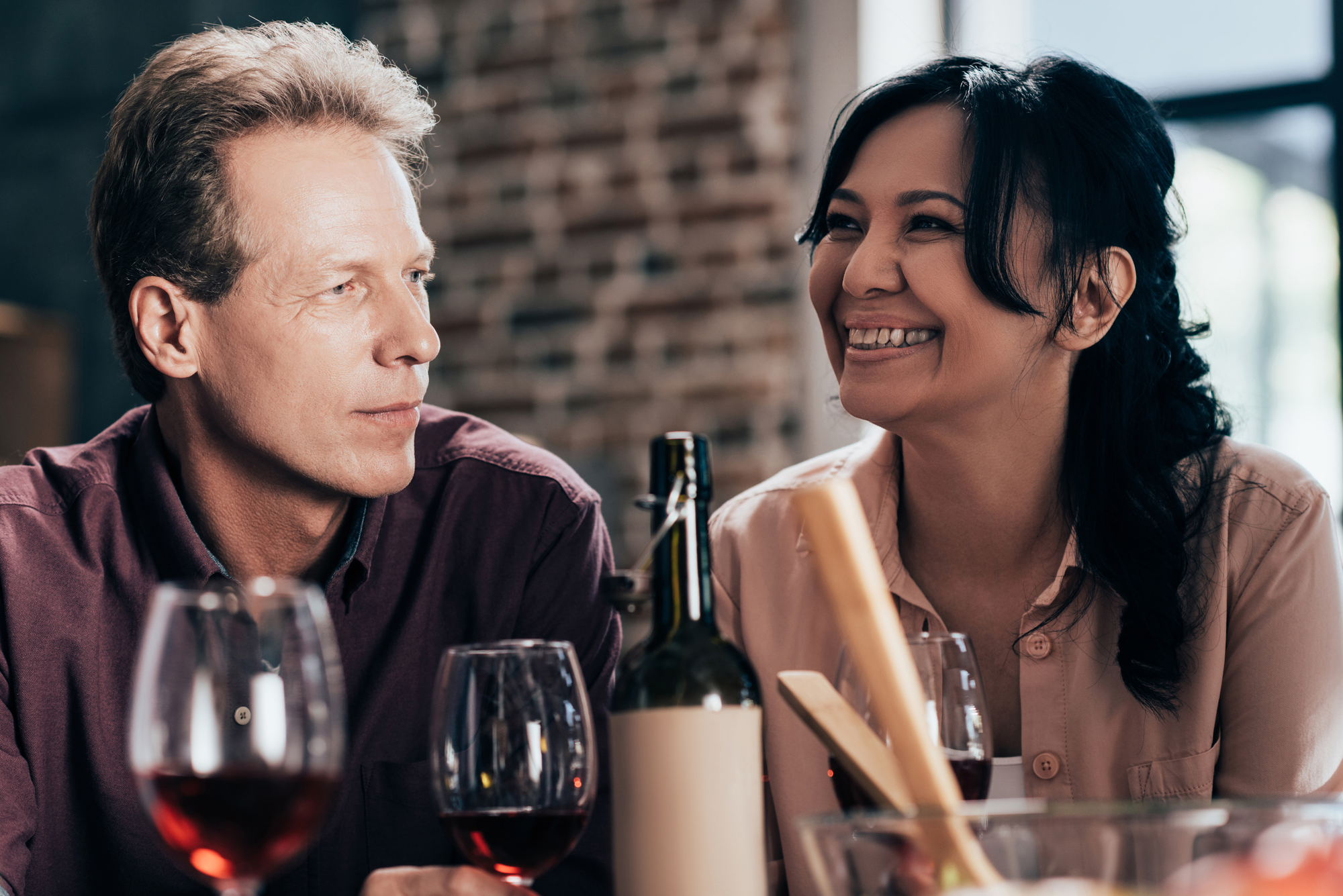 A man and a woman are sitting at a table with wine glasses and a wine bottle in front of them. The woman is smiling brightly, and the man is looking at her with a slight smile. They appear to be enjoying a pleasant moment together in a cozy, brick-walled setting.