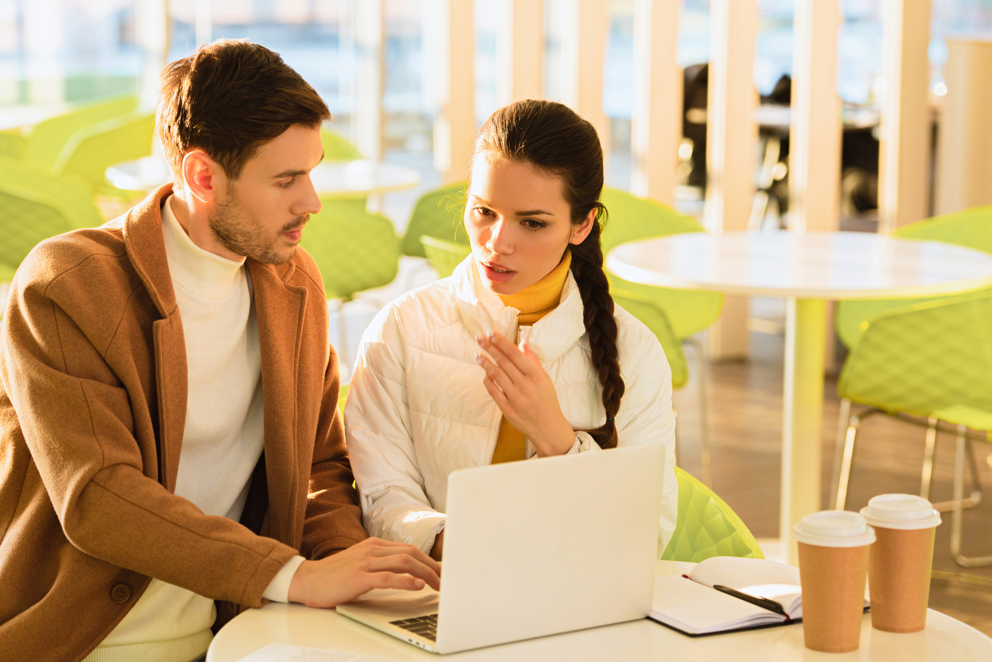 A man and a woman are sitting at a table collaborating over a laptop in a bright, modern cafe. The woman is gesturing as she speaks, while the man, wearing a brown coat, types on the keyboard. There are two coffee cups and an open notebook on the table.