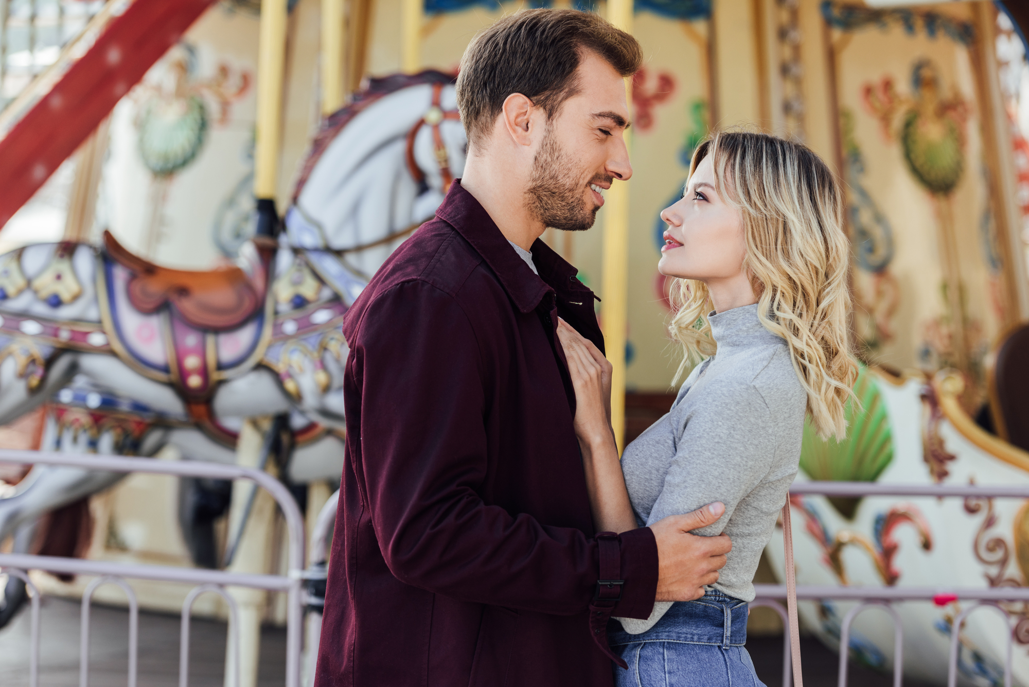 A couple stands facing each other in front of a colorful carousel, with the woman resting a hand on the man's chest. The man wears a burgundy jacket, and the woman wears a gray long-sleeve top and blue jeans. Both are gazing into each other's eyes.
