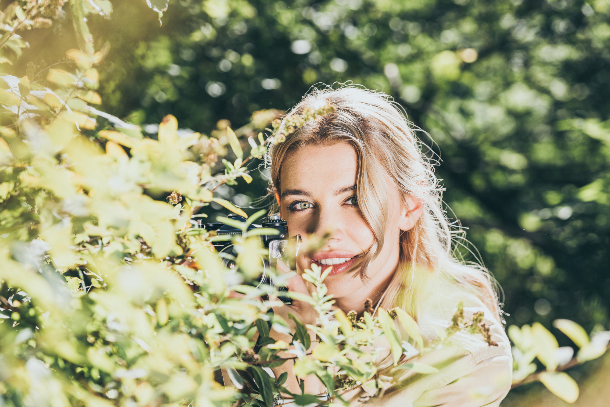 A smiling woman with blonde hair is partially hidden behind green leaves, enjoying a sunny day outdoors. She is looking at the camera with bright blue eyes. The background is filled with lush, green foliage, creating a vibrant and fresh atmosphere.