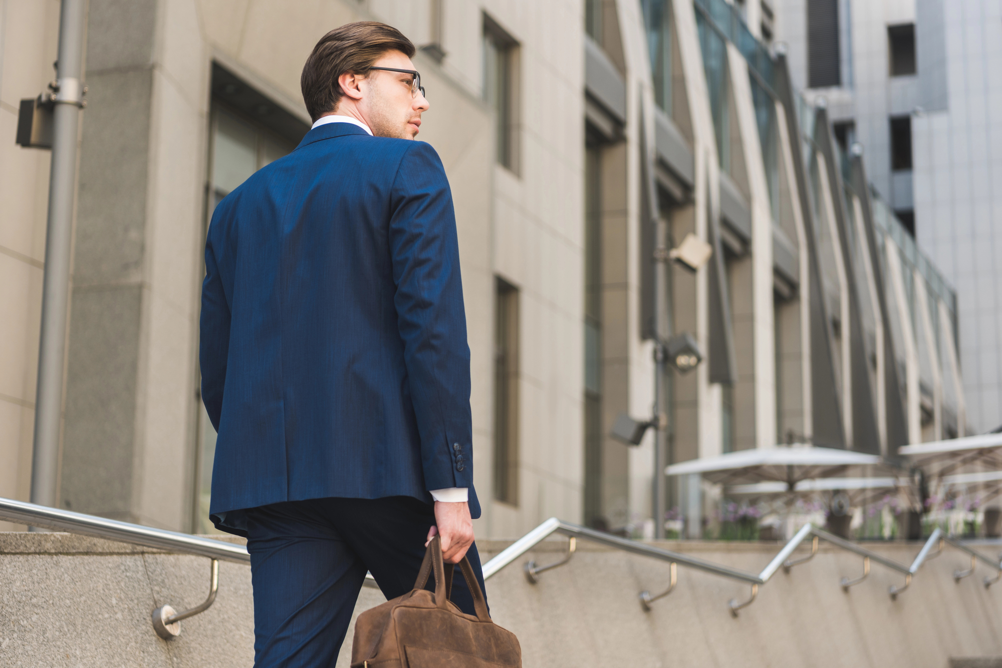 A man dressed in a navy blue suit and sunglasses walks up a set of exterior stairs with a brown leather briefcase in his hand. He is surrounded by modern office buildings, and the scene is set during the day.