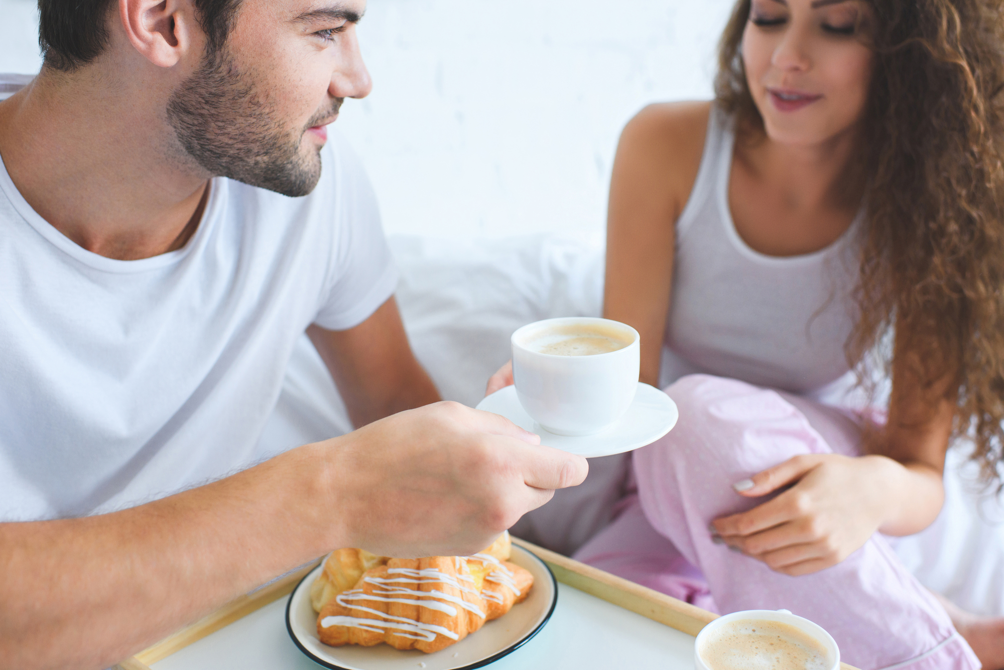 A man and woman in casual clothing sit closely together, enjoying a breakfast in bed. The man holds a cup of coffee on a saucer towards the woman, who is smiling. A tray with a croissant is placed on the bed in front of them.