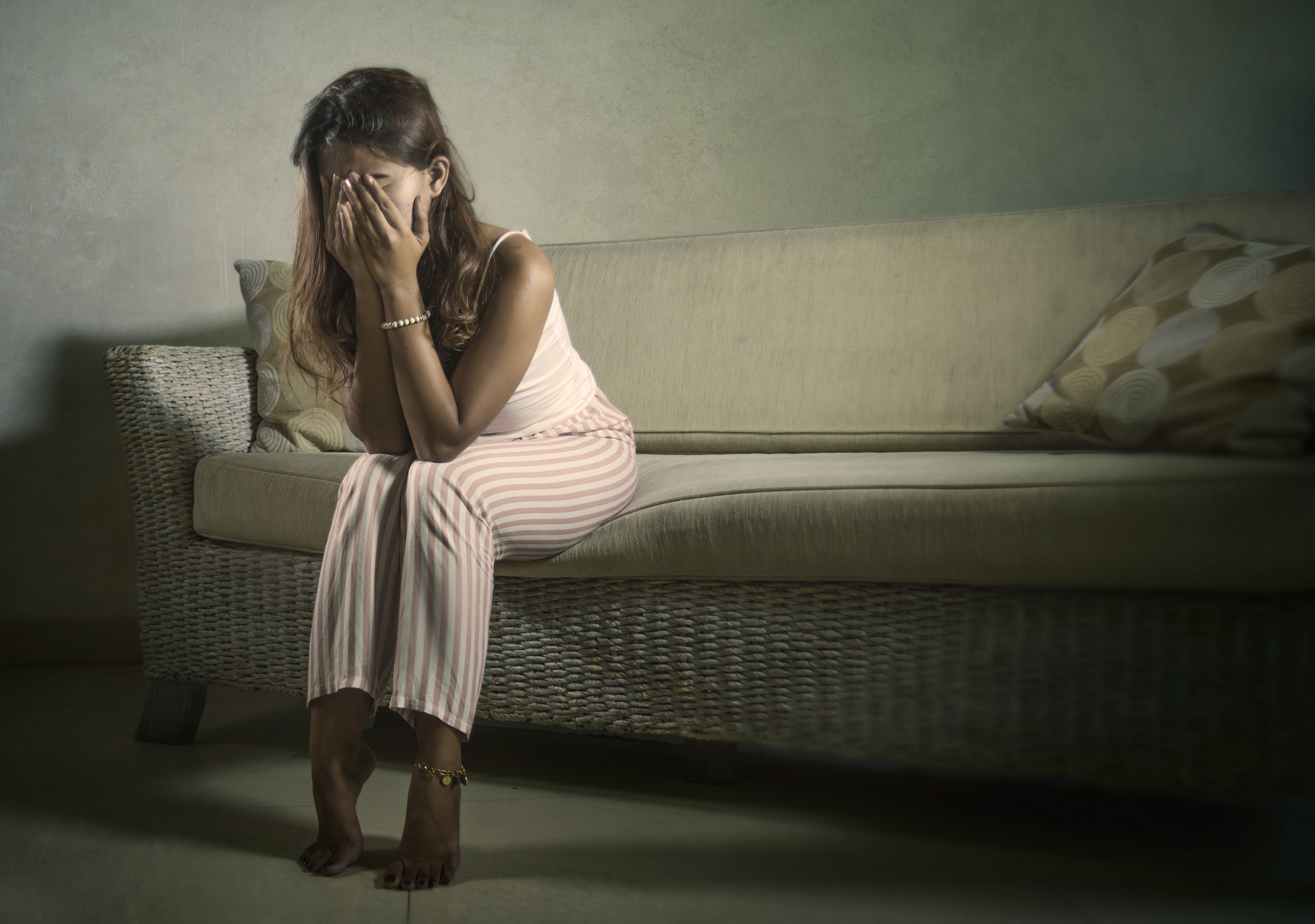 A woman sits on a cushioned, wicker sofa with her hands covering her face. She appears distressed, wearing a sleeveless top and striped pants. The room has a neutral-toned, textured wall with a patterned pillow on the sofa, creating a subdued atmosphere.