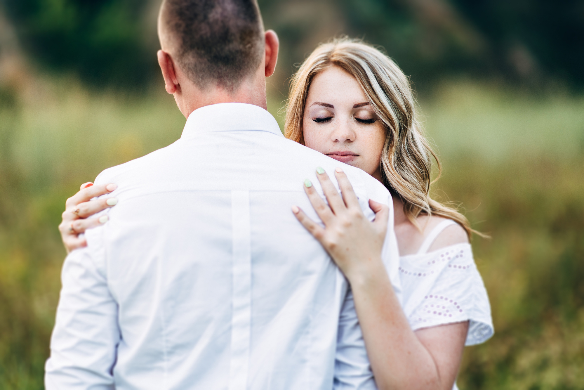 A woman in a white dress embraces a man in a white shirt, resting her head against his back with her eyes closed. They stand outdoors in a grassy field, with a blurred natural background.