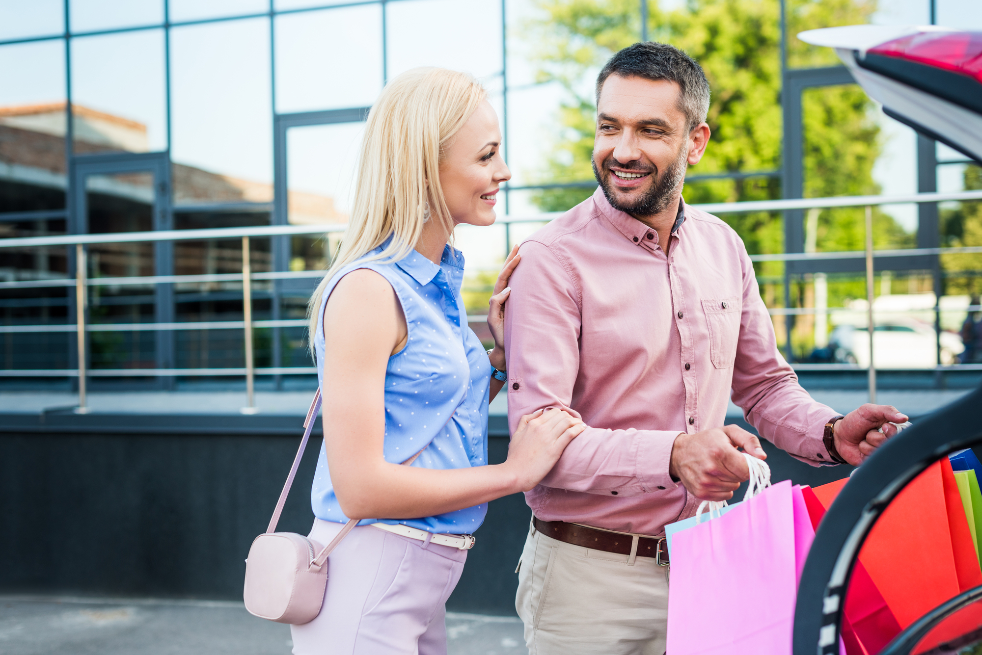 A smiling couple stands beside an open car trunk outside a modern building with reflective windows. The man, holding colorful shopping bags, looks at the woman, who has her hand on his arm. Both are casually dressed and seem to be enjoying their time together.