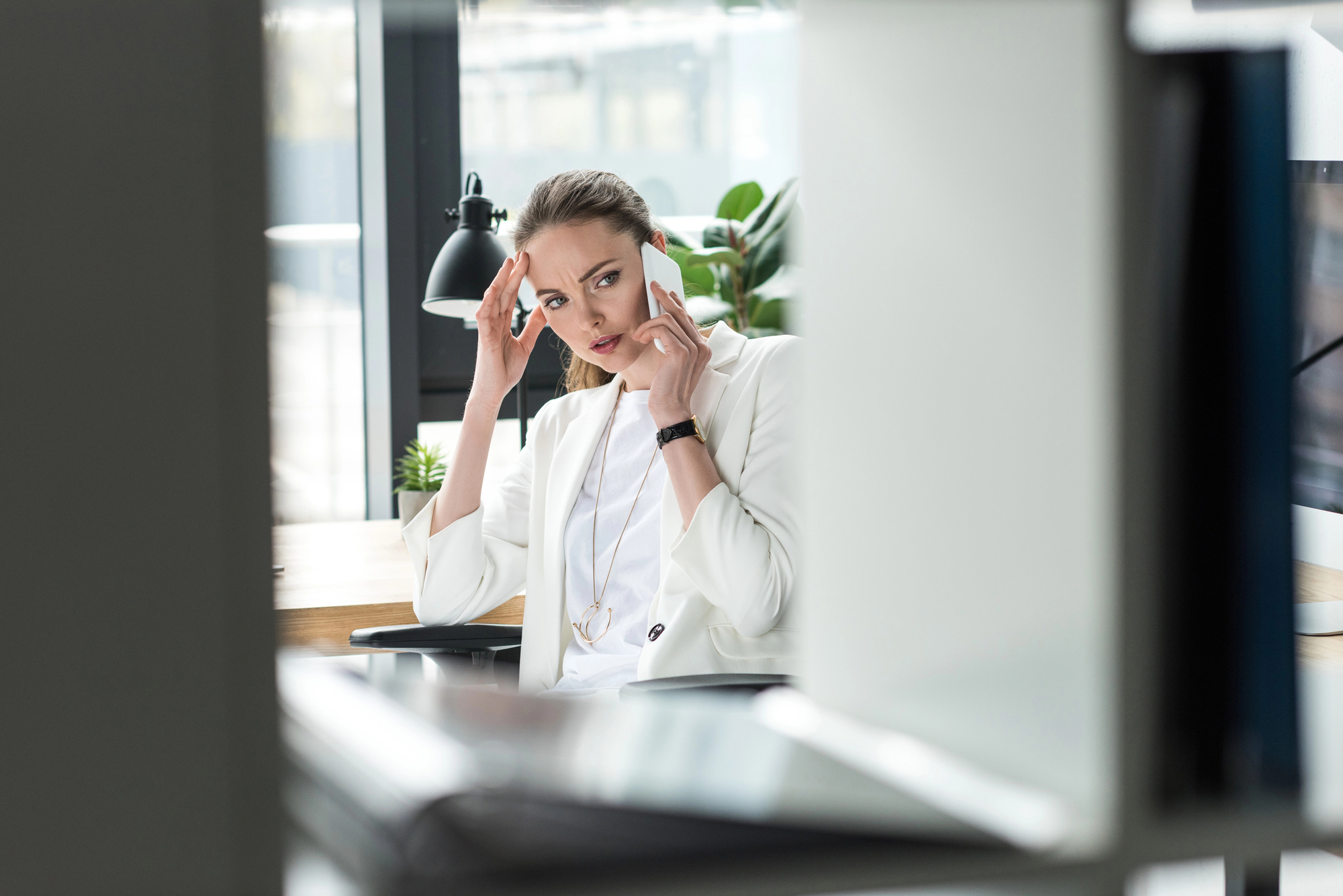 A woman dressed in white sits at a desk in an office, holding her temples and looking stressed. She has long, brown hair tied back, and the desk features a lamp and plant. The background includes large windows and a computer monitor.