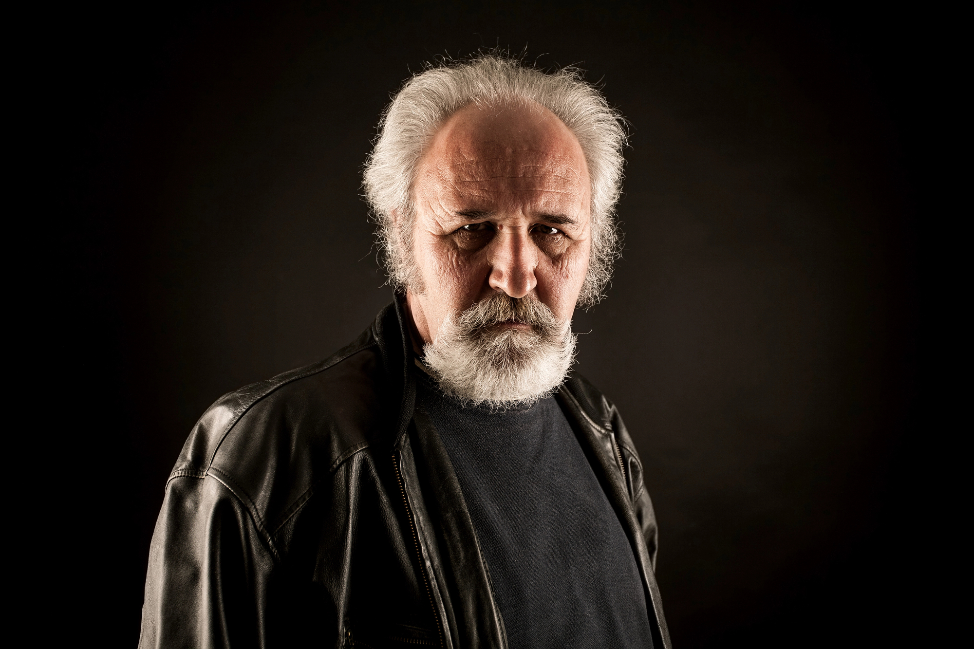 A mature man with a serious expression, gray hair, and a full white beard stands against a dark background. He is wearing a black leather jacket and a black shirt. His intense gaze is directed at the camera.
