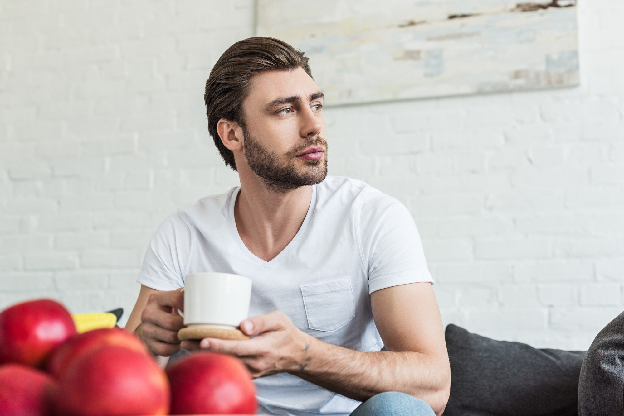 A man with brown hair and a beard, wearing a white t-shirt, sits on a couch and holds a white mug with both hands. In the foreground, there are several red apples on a table. The background features a white brick wall and a blurred painting.