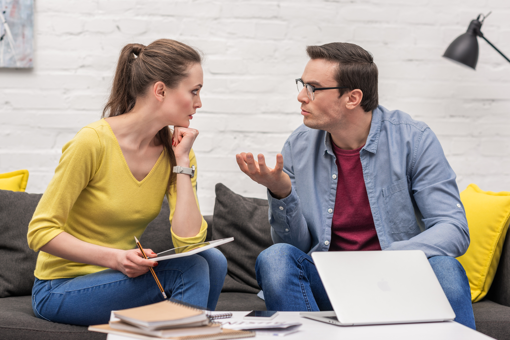 A woman and a man sit on a sofa having a serious conversation. The woman, wearing a yellow sweater, holds a notebook and pencil, while the man, in a blue shirt and glasses, gestures with his hand. A laptop and notebooks are on the table in front of them.