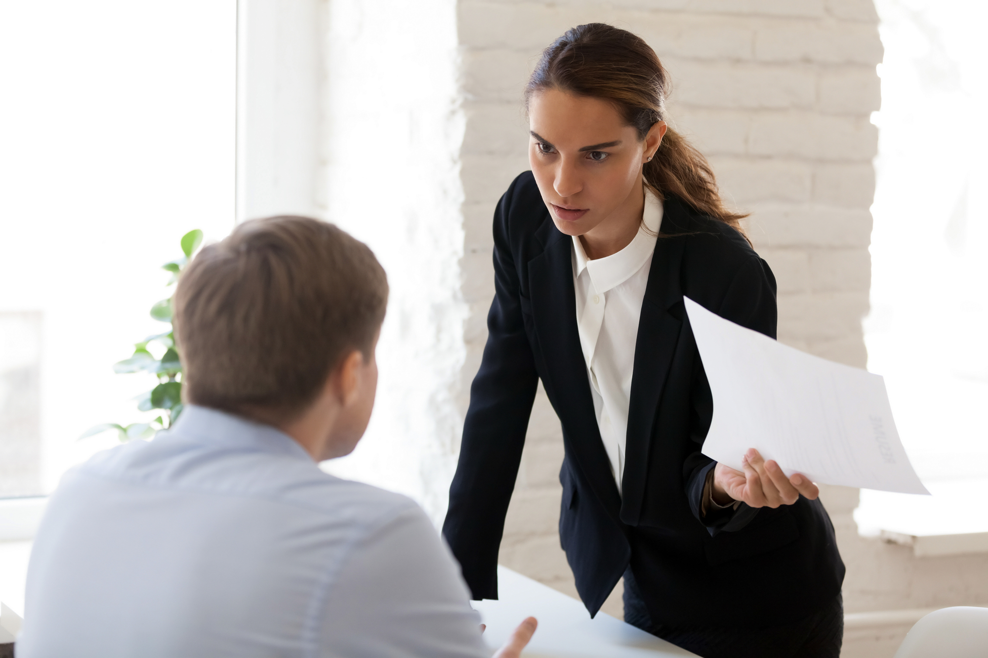A woman in a formal black blazer and white shirt holds a piece of paper while leaning towards a man sitting at a desk, seemingly in a serious conversation. The man, dressed in a light blue shirt, sits facing her. They are in a brightly lit office.