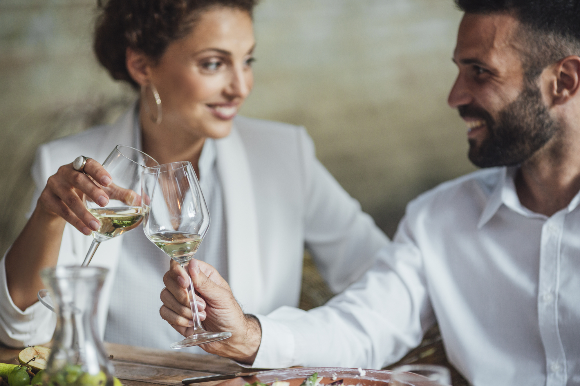 A woman and a man, both dressed in white outfits, are seated at a table and clinking wine glasses together, smiling warmly at each other. The table has some food and a decanter of wine. The background is softly blurred.