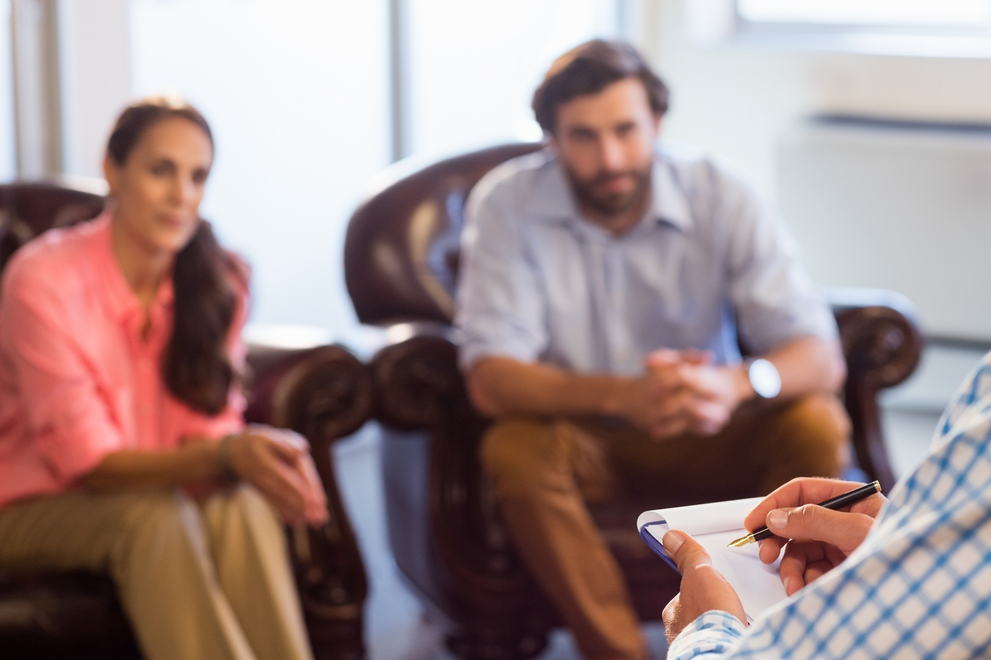 Two people sit on a couch, facing a person holding a notepad and pen. The person with the notepad is presumably leading a discussion or counseling session. The room is well-lit with natural light streaming through windows in the background.