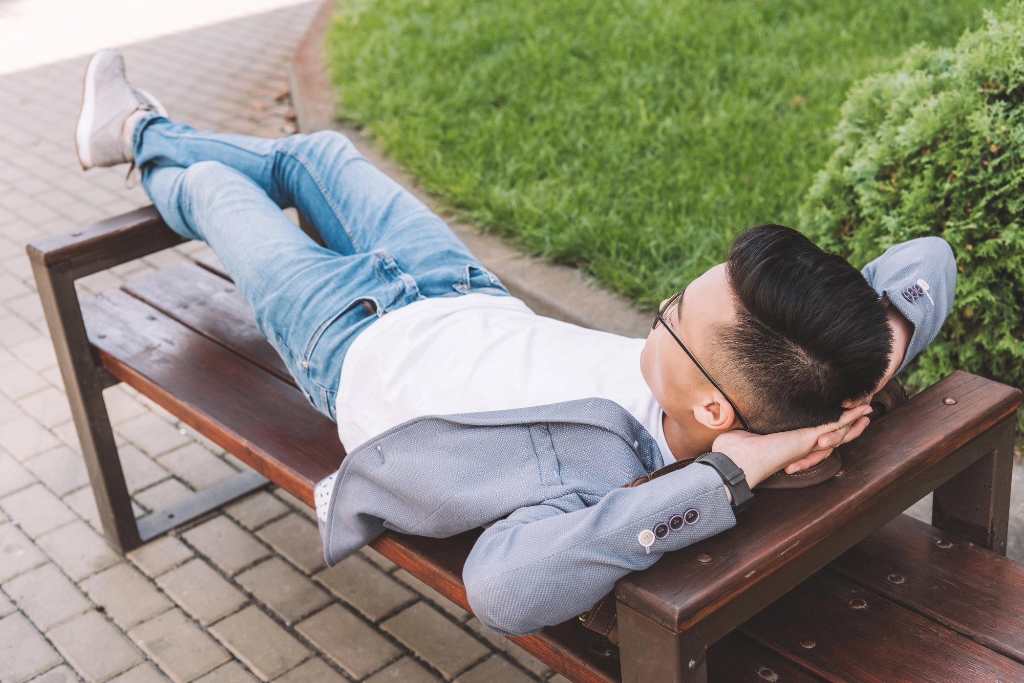 A person wearing glasses, a gray blazer, white shirt, jeans, and white shoes is lying on a wooden bench, with hands behind their head and legs crossed, relaxing outdoors on a sunny day. Green grass and a paved walkway are seen in the background.