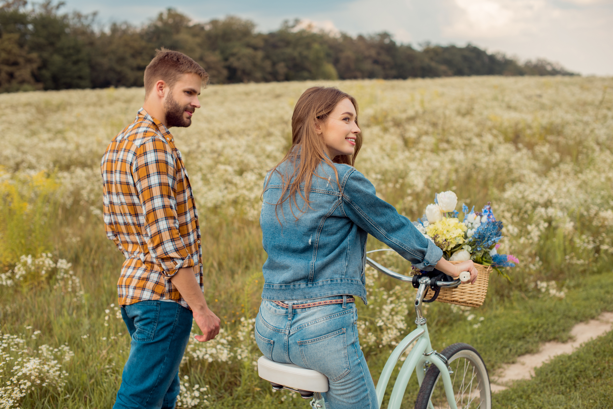 A man and woman are enjoying a day outdoors in a scenic field of wildflowers. The woman, wearing a denim jacket, is on a bicycle with a basket full of flowers, while the man in a plaid shirt walks beside her. The sky is partly cloudy, and trees fill the background.