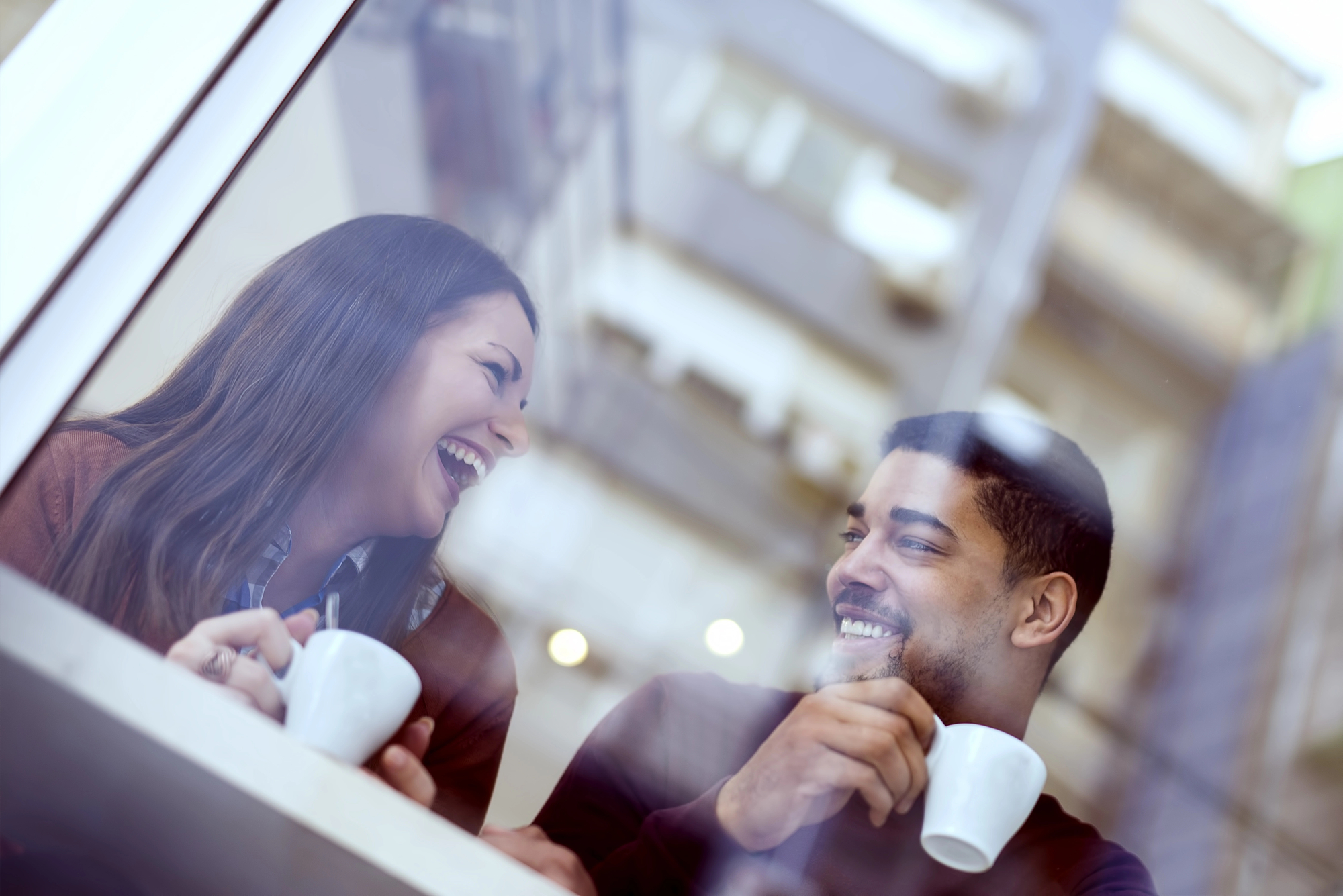 Two people sit at a café table, each holding a coffee cup and smiling warmly at each other. The image is viewed through a window, with soft reflections of lights and surrounding buildings. Both individuals appear to be enjoying their conversation.
