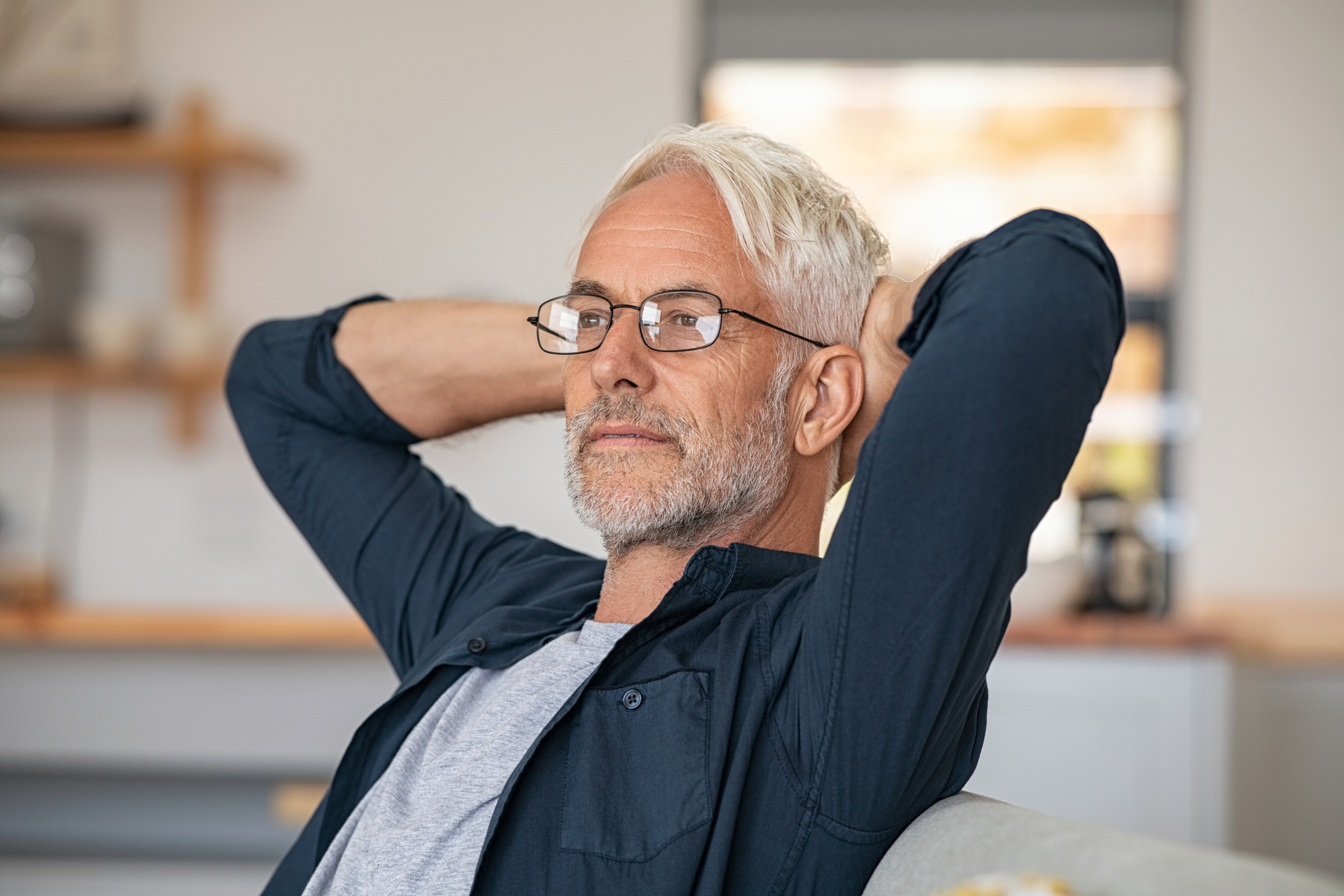 A mature man with gray hair and beard, wearing glasses and a dark shirt, is seated indoors. He has his hands behind his head, looking relaxed and pensive. The background shows blurred kitchen shelves and a window.