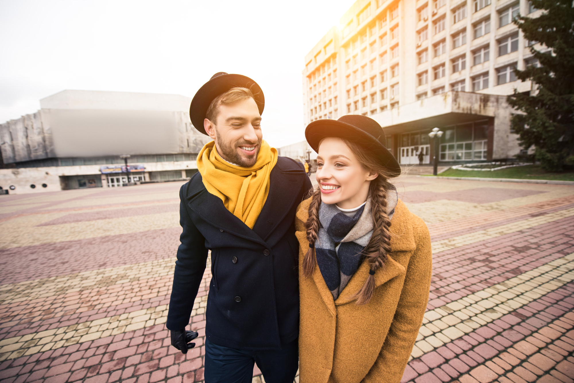 A young couple walks cheerfully through a paved square on a sunny day. Both are wearing hats and cozy winter coats with scarves. The man has a black coat and yellow scarf, while the woman wears a brown coat and gray scarf. Buildings stand in the background.