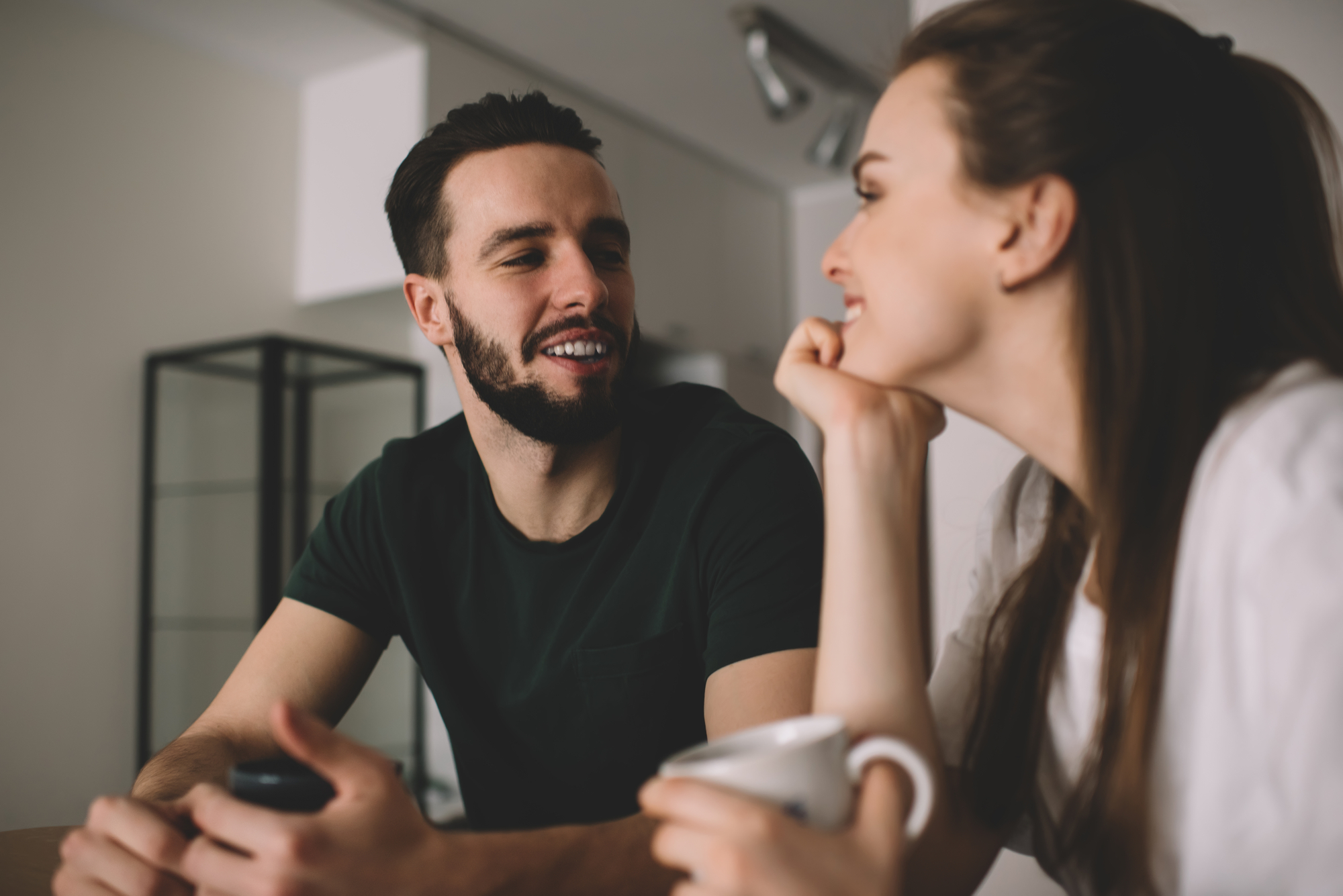 A man with a beard and a woman with long hair sit at a table, smiling at each other. The man is holding a smartphone while the woman is holding a white coffee mug. They are in a well-lit, modern room with minimalistic decor.