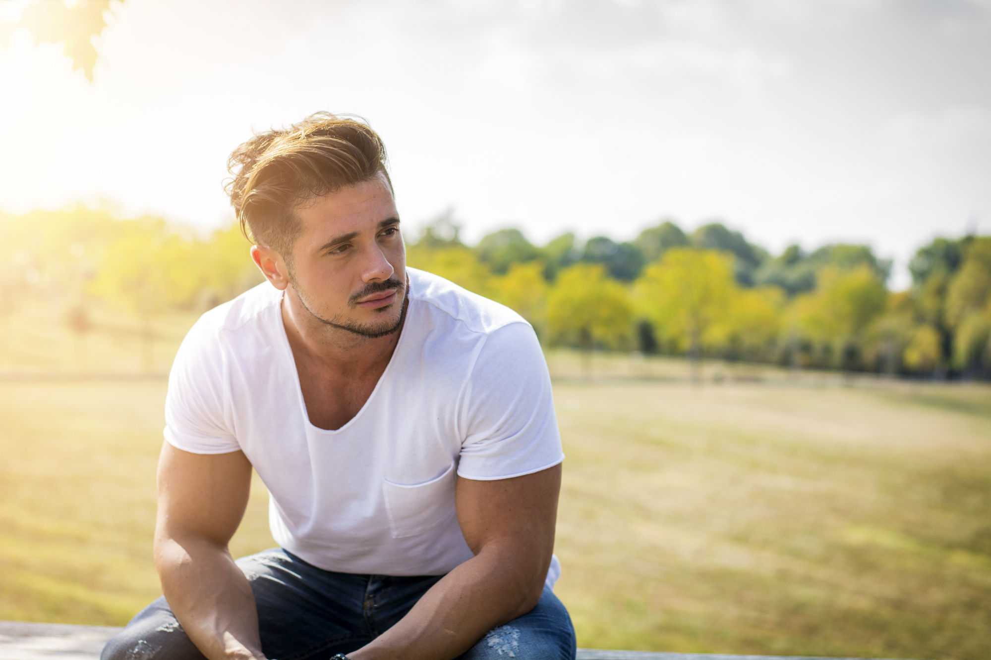 A man with short hair sits on a bench outdoors in a sunny park. He is wearing a white T-shirt and blue jeans, looking pensively into the distance. Green trees and soft sunlight create a peaceful background.
