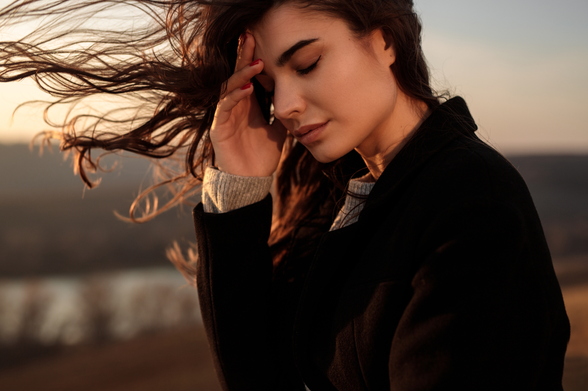 A woman with long, flowing hair stands outdoors with her eyes closed, gently touching her forehead. She is dressed in a black coat. The blurry background shows a serene landscape at sunset. Her expression is peaceful and contemplative.
