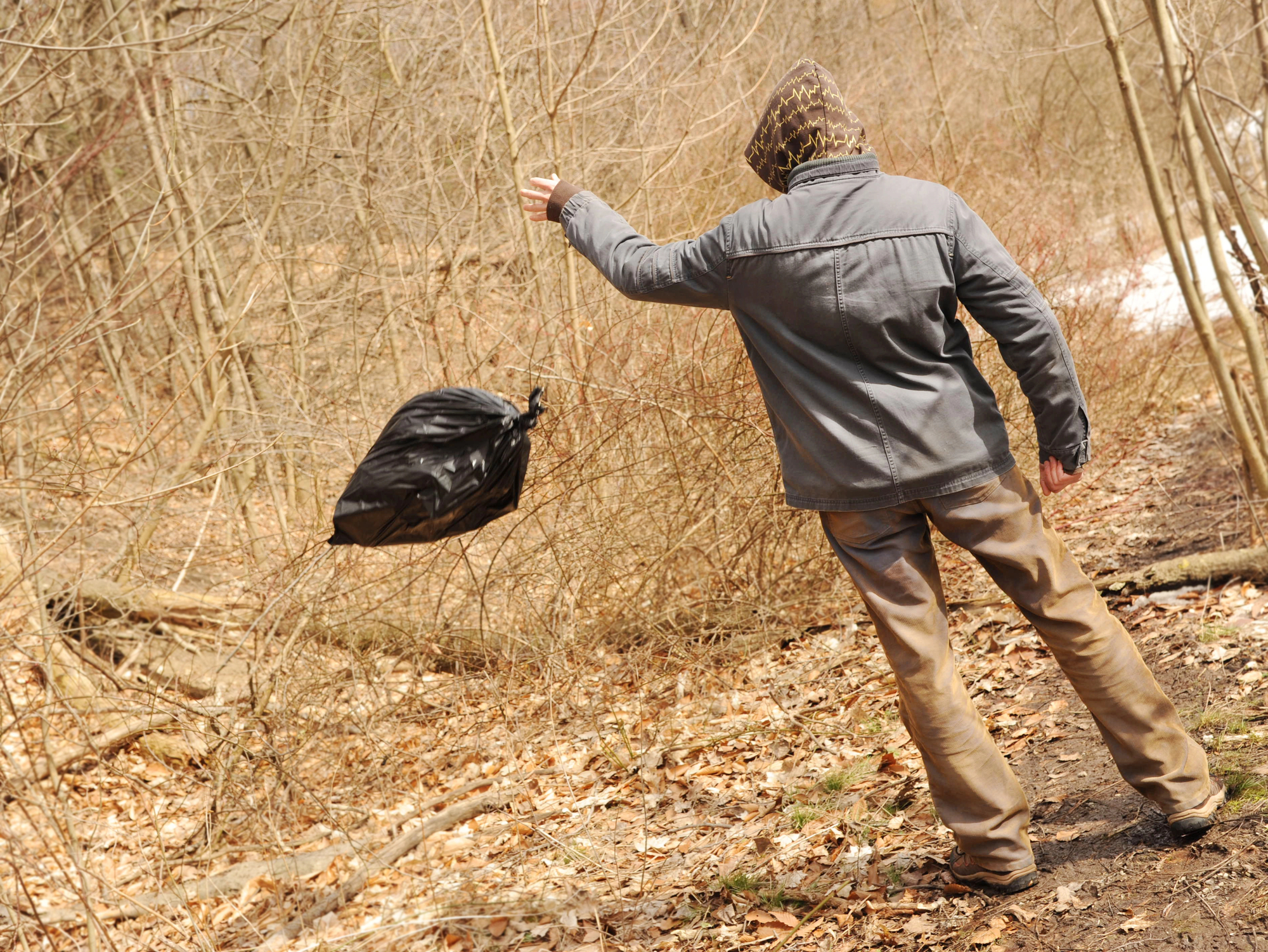 A person in a gray jacket and camouflage hood throws a black garbage bag into a forest area with leafless trees and branches. The ground is covered with dry leaves. The scene depicts littering in nature.