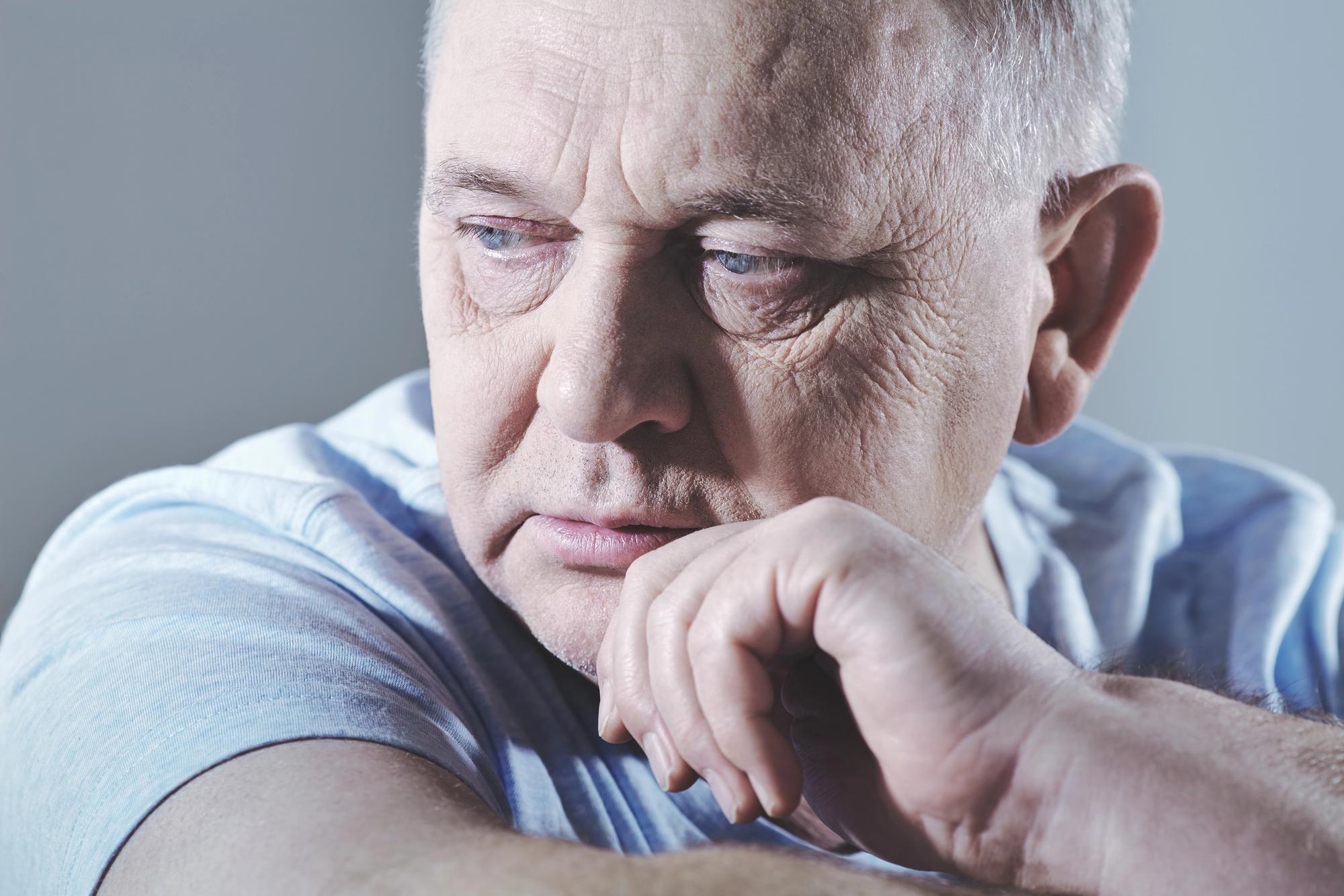 An elderly man with a worried, pensive expression rests his chin on his hand. He has short hair and is wearing a light-colored shirt. He looks off to the side, and the background is a neutral, soft-focus gray.