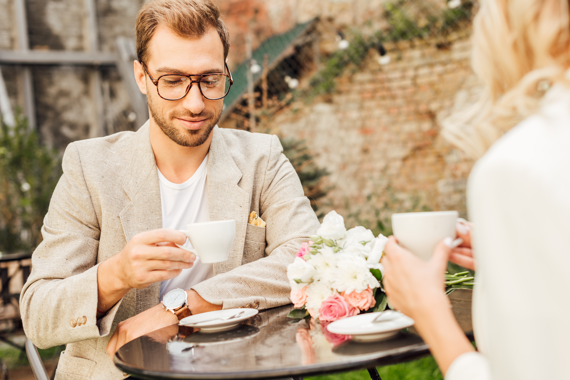 A man with glasses and a beard wearing a beige blazer is sitting at a cafe table outdoors, holding a cup of coffee. Across from him is a woman with blonde hair holding a cup. The table is adorned with a bouquet of flowers. A stone wall and string lights are in the background.