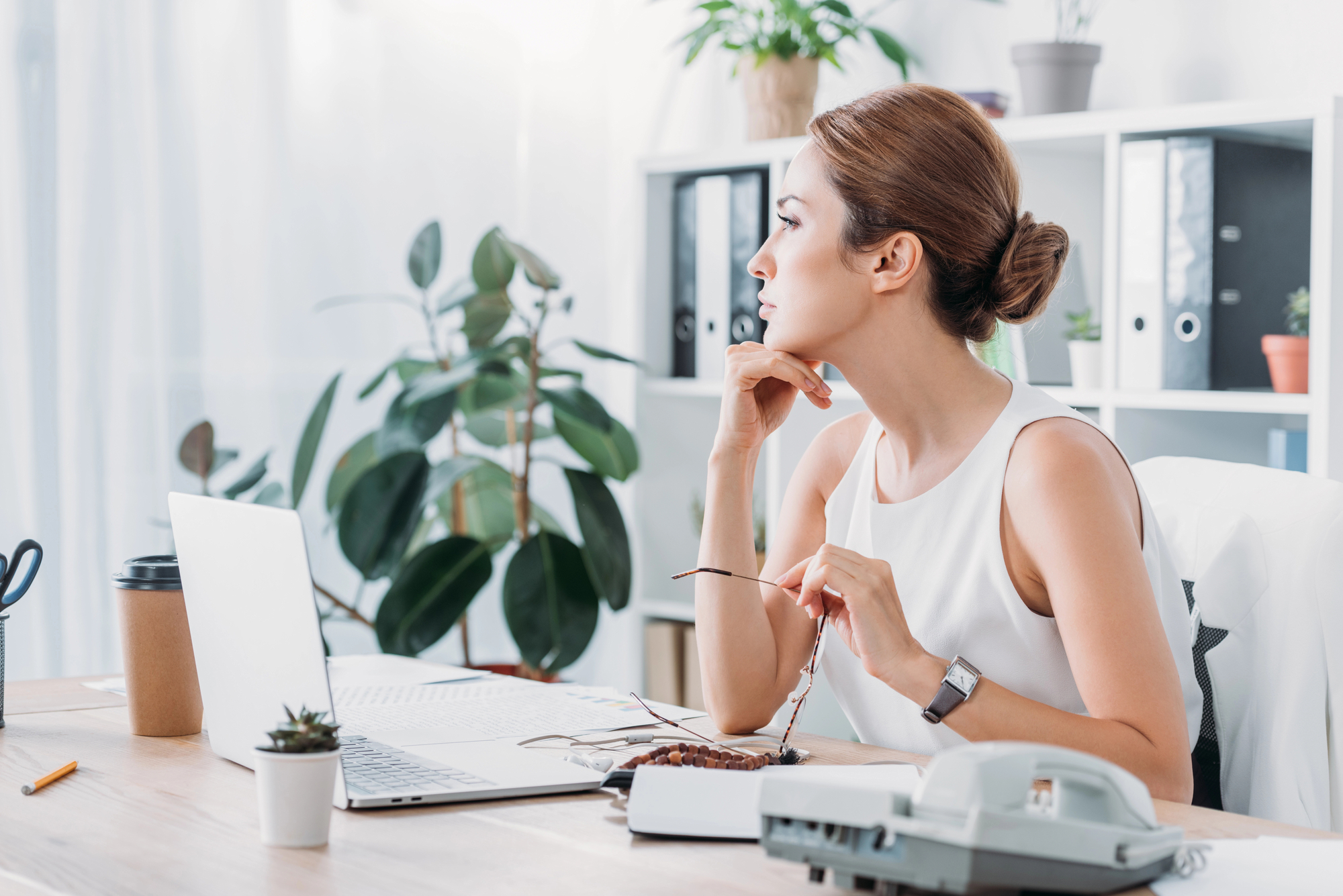 A woman sits at a desk in an office, looking thoughtfully out the window. She holds a pair of glasses in one hand and has a laptop, a coffee cup, a potted plant, and office supplies on the desk. Shelves with folders and plants are visible in the background.