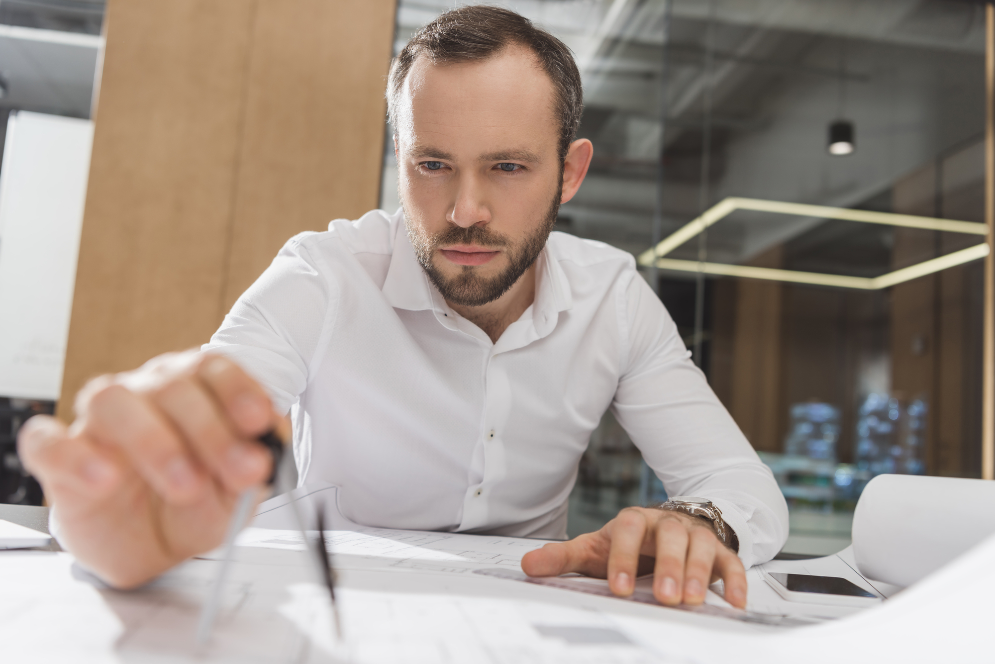 A man in a white shirt is intently working at a desk, holding a compass over architectural plans or blueprints. He is focused and appears to be in an office with glass walls and modern lighting in the background.