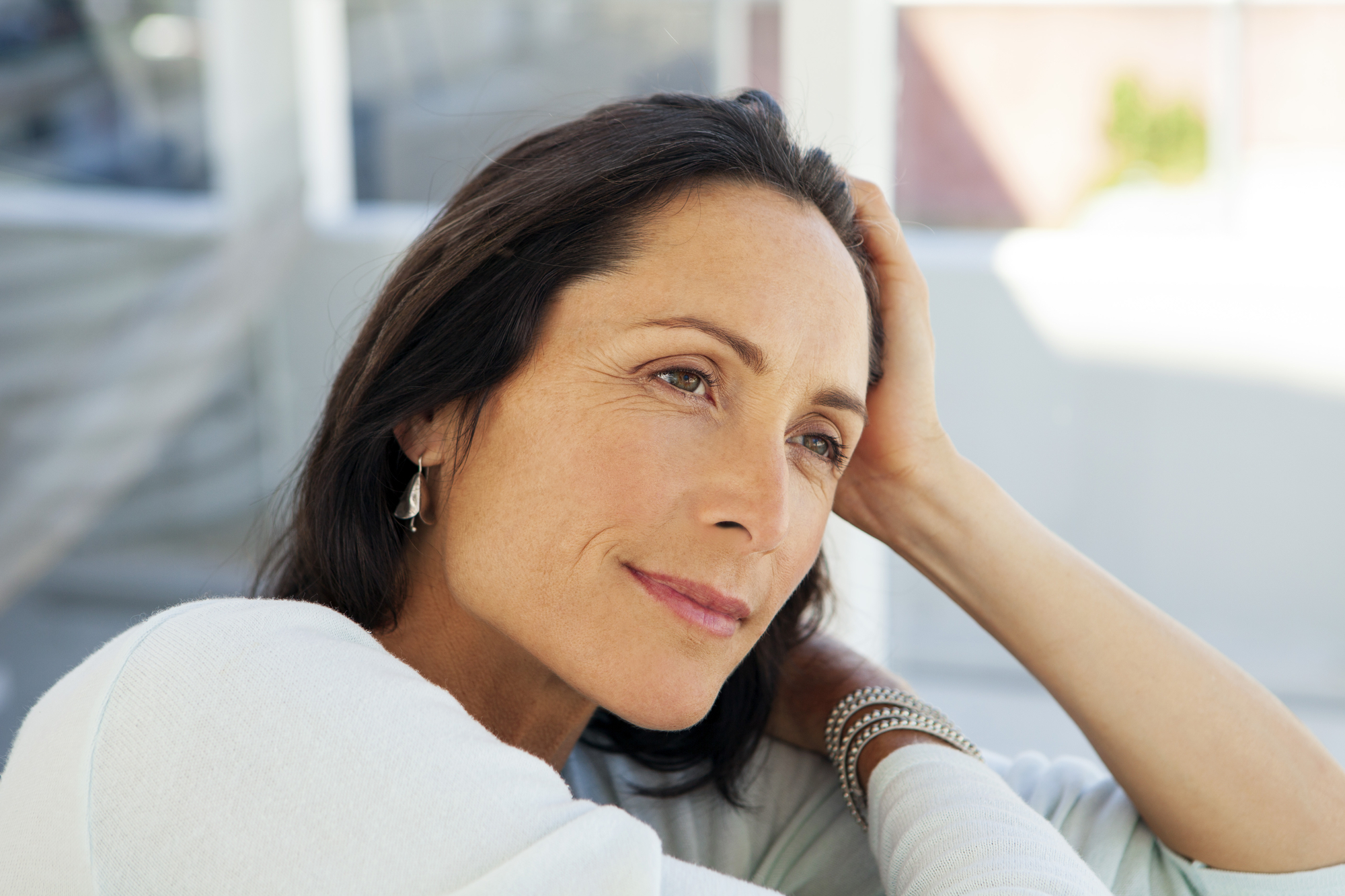 A woman with long dark hair leans her head on her hand, gazing thoughtfully into the distance. She is wearing a light-colored top and silver earrings, with bracelets on her wrist. The background is blurred, suggesting a peaceful, sunlit setting.