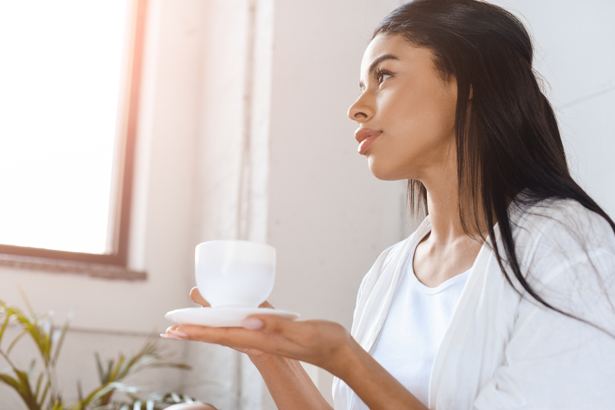 A woman with long dark hair wearing a white shirt is holding a white cup and saucer. She is gazing out of the window, bathed in soft natural light. In the background, there is a green plant and a white wall.
